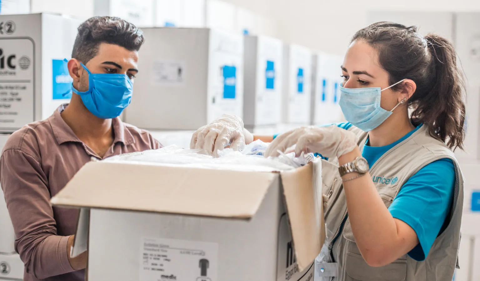 A man and a woman pack up a box of vaccines. They are both wearing masks and the woman is wearing a UNICEF vest.