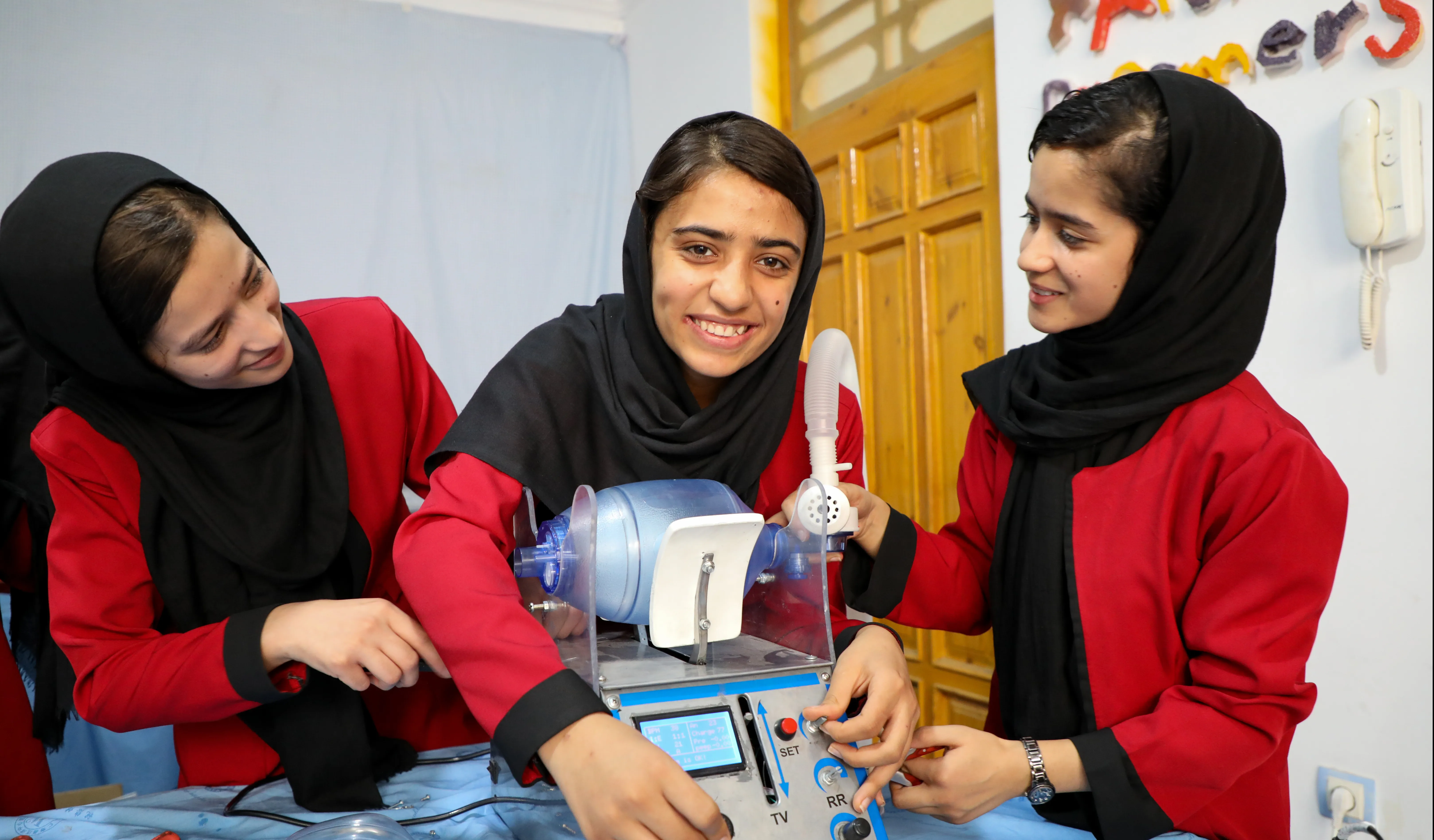 Three girls in red uniforms and black headscarves stand around a desk with a ventilator on it. 
