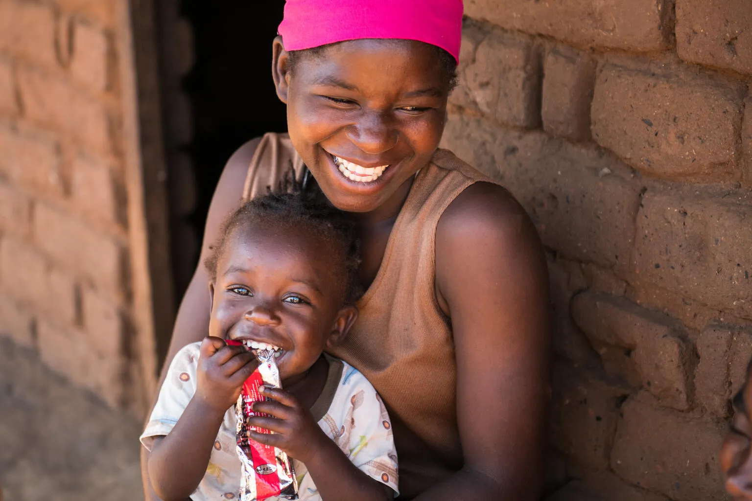 A smiling woman embraces a giggling child holding a packet of peanut paste. 