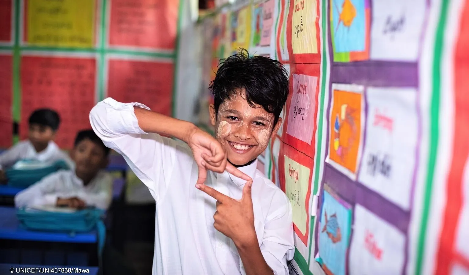 A boy uses his hands to create a square photo sign. He is in a classroom setting standing next to bulletin board with many colourful sheets pinned on it. He is wearing a white uniform shirt. In Cox's Bazaar, Bangladesh. 