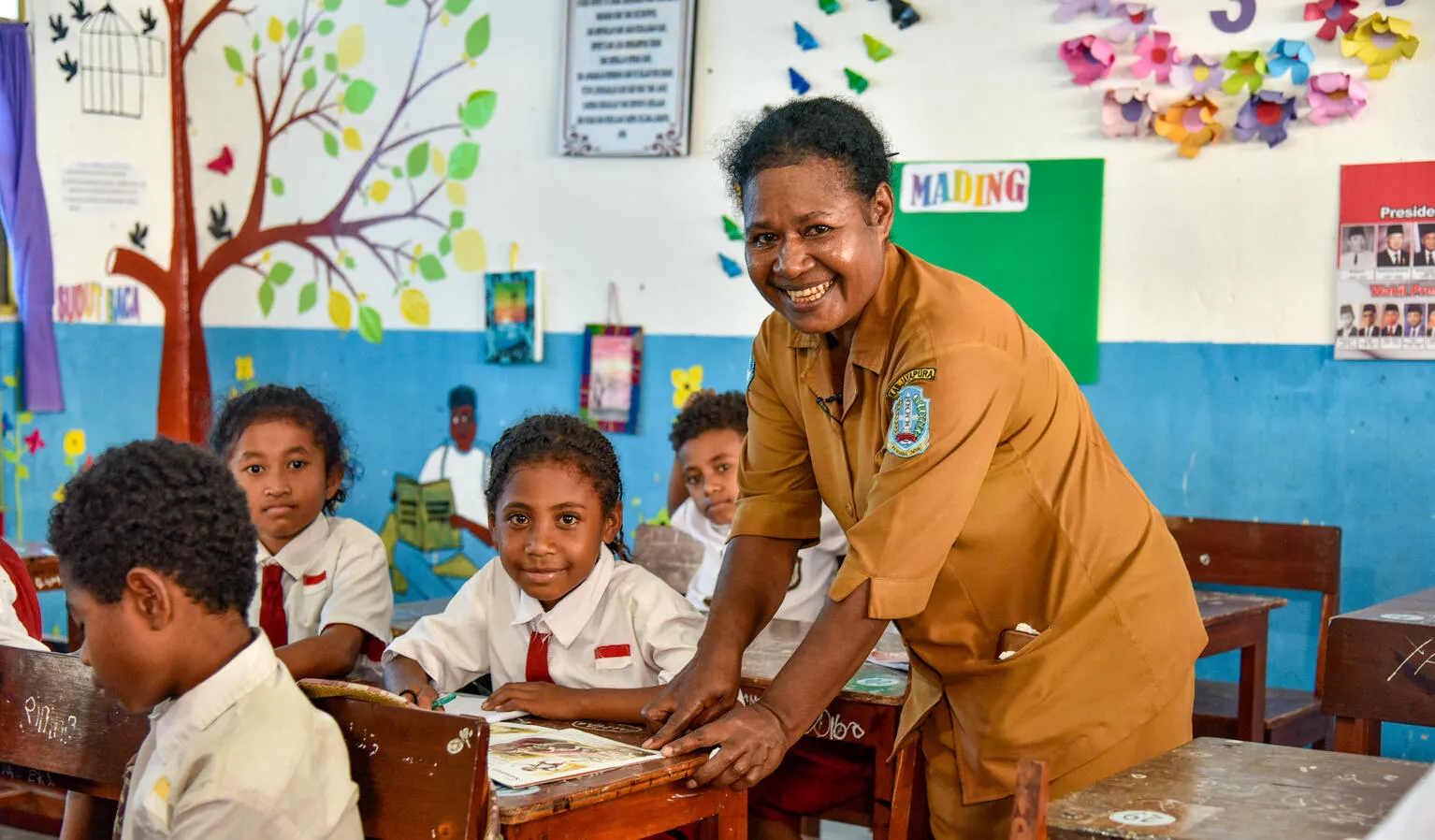 A teacher leans over a students desk and smiles at the camera. The student next to her is also smiling. Photo taken in Indonesia.