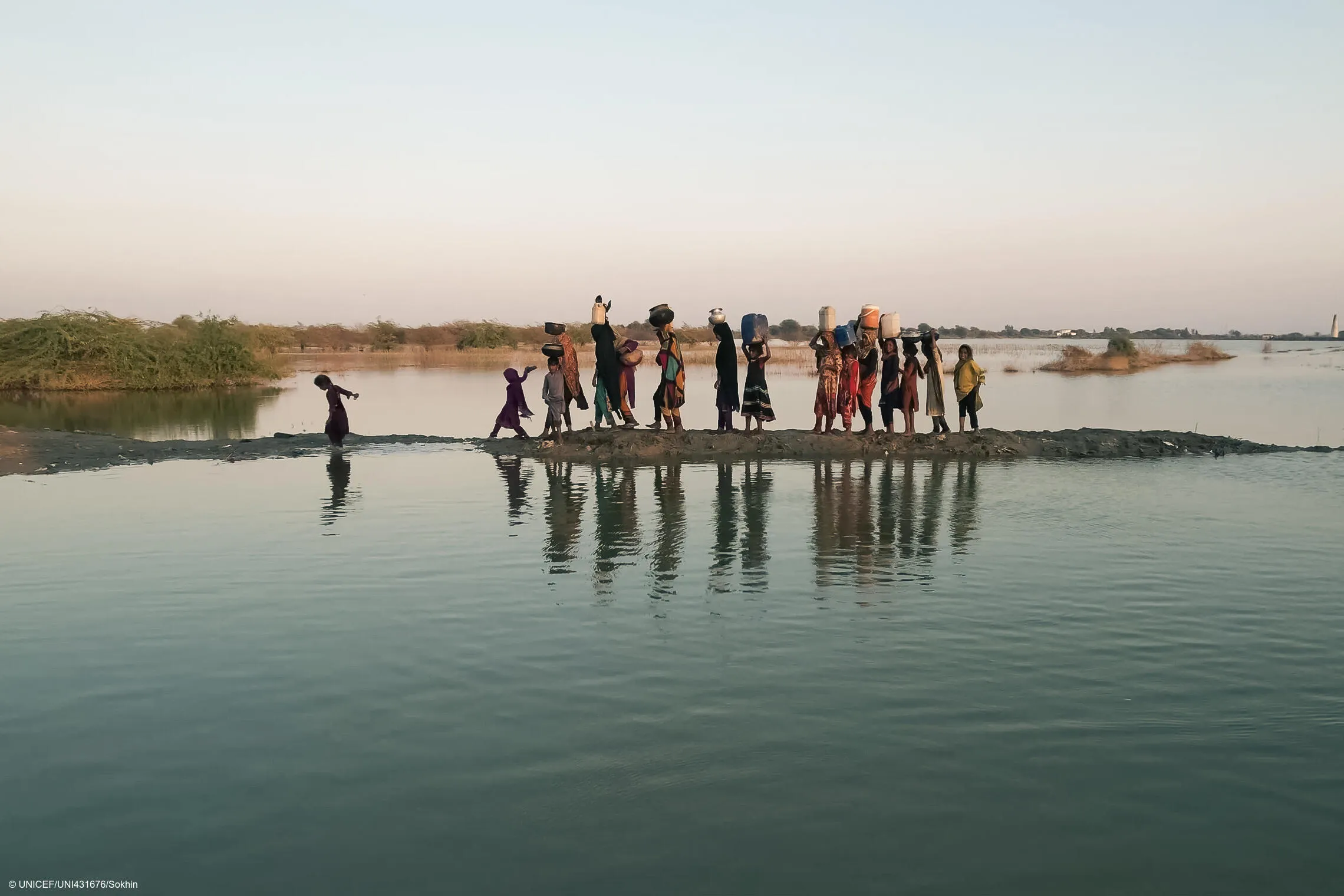 A group of people, including women and children, walk with water containers on their heads along a narrow, muddy path flanked by water in a rural setting.