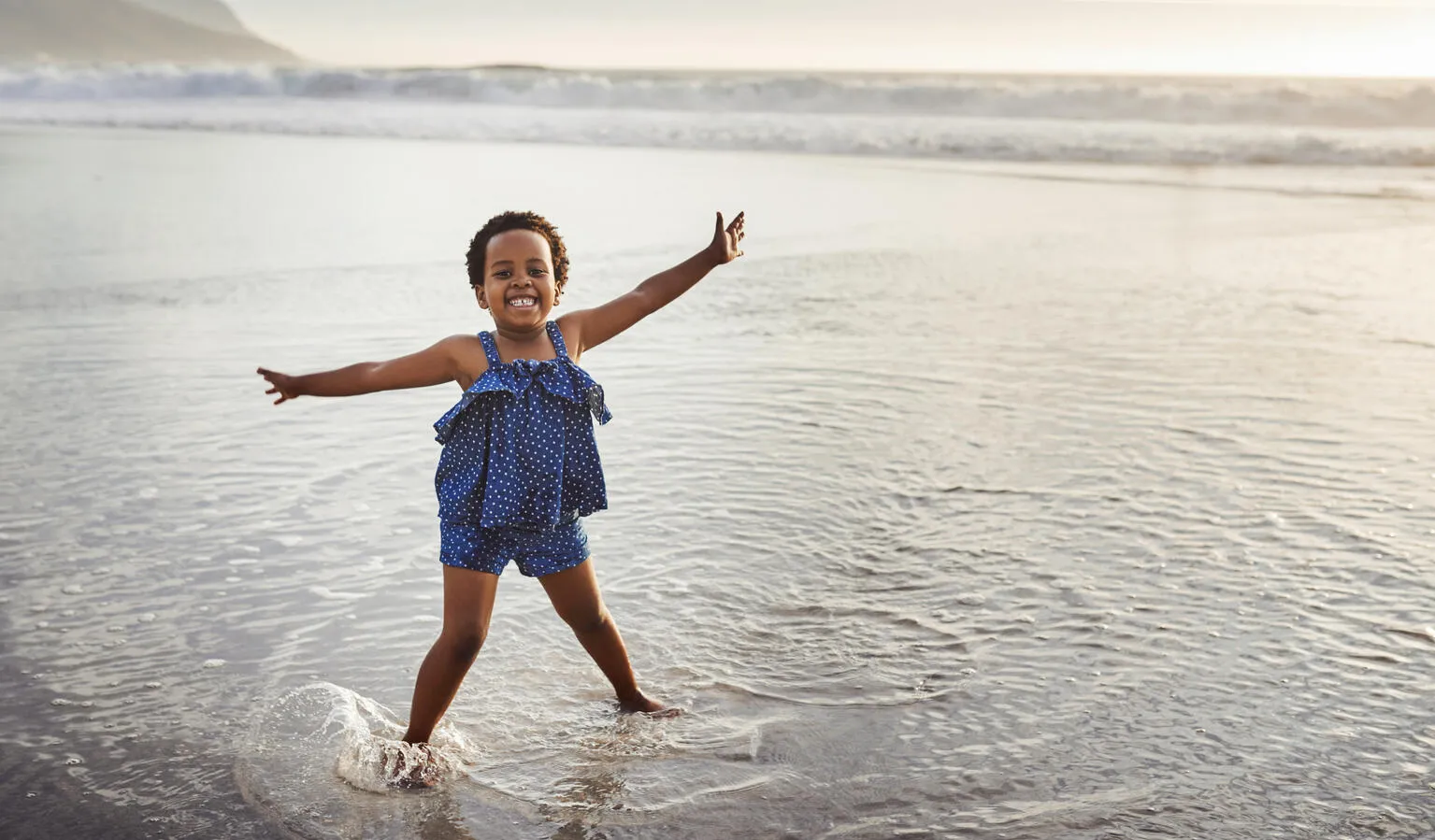 A young girl in stands in a body of water with her arms held wide smiling at the camera as the sun sets to the side of the photo