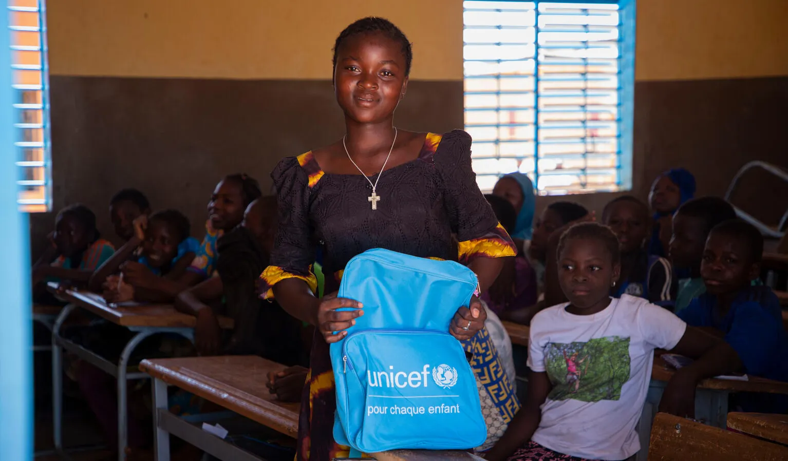 A girl hold a blue UNICEF backpack in front of a classroom full of students.