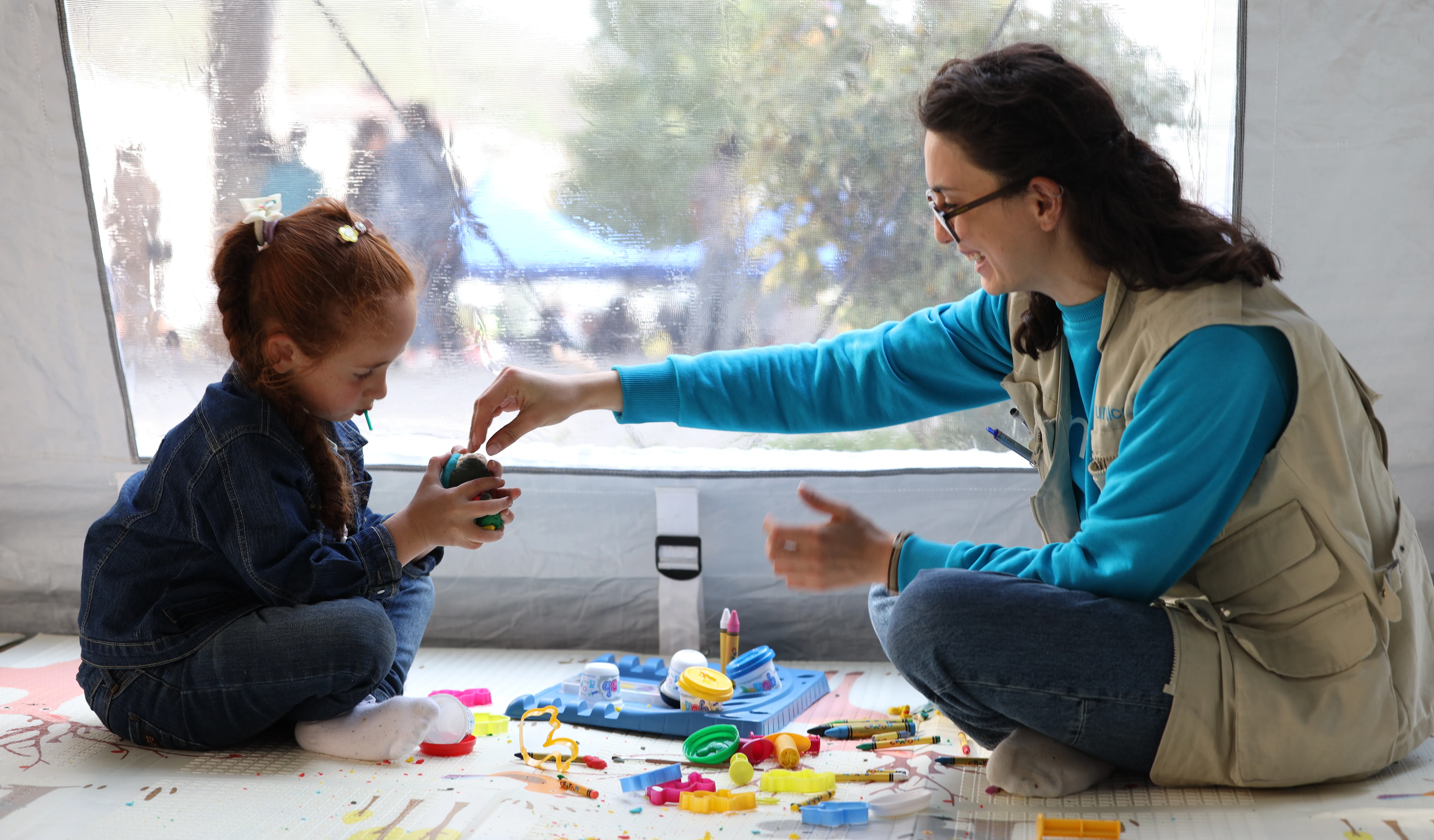 A child and a UNICEF staff member sit in a tent on the floor. The staff member giving the child a block with other toys lying around them.