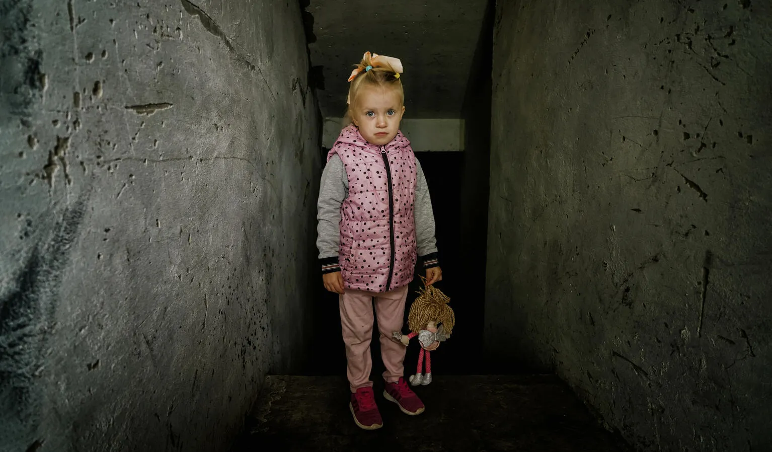 A young girl stands in a dimly lit, narrow concrete passage. She wears a pink polka-dotted vest, holding a stuffed toy, with a somber expression.