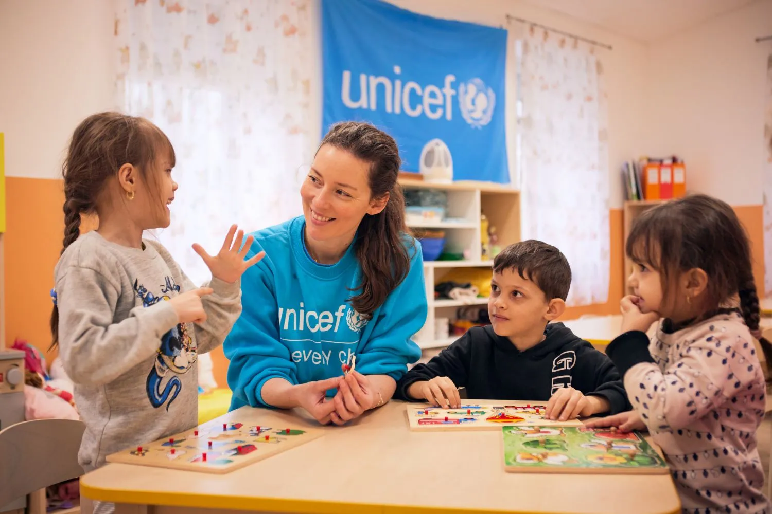 Two girls and a boy play around a table with a woman wearing a blue UNICEF sweater. 