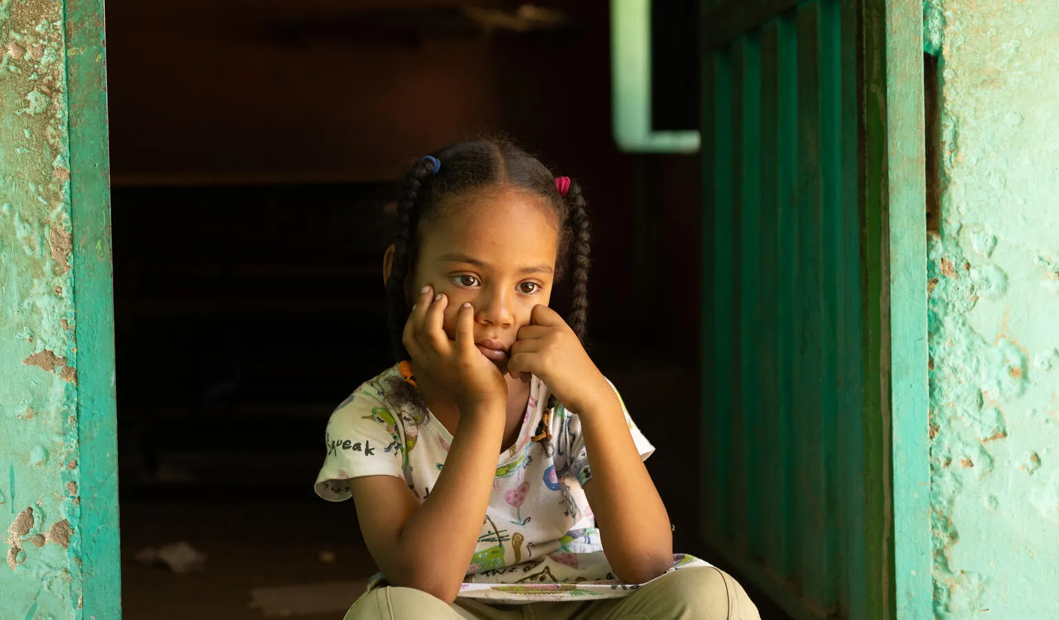 Young girl with pigtails, wearing a patterned shirt, sits pensively in a dimly lit doorway. She rests her chin on her hands, appearing thoughtful.