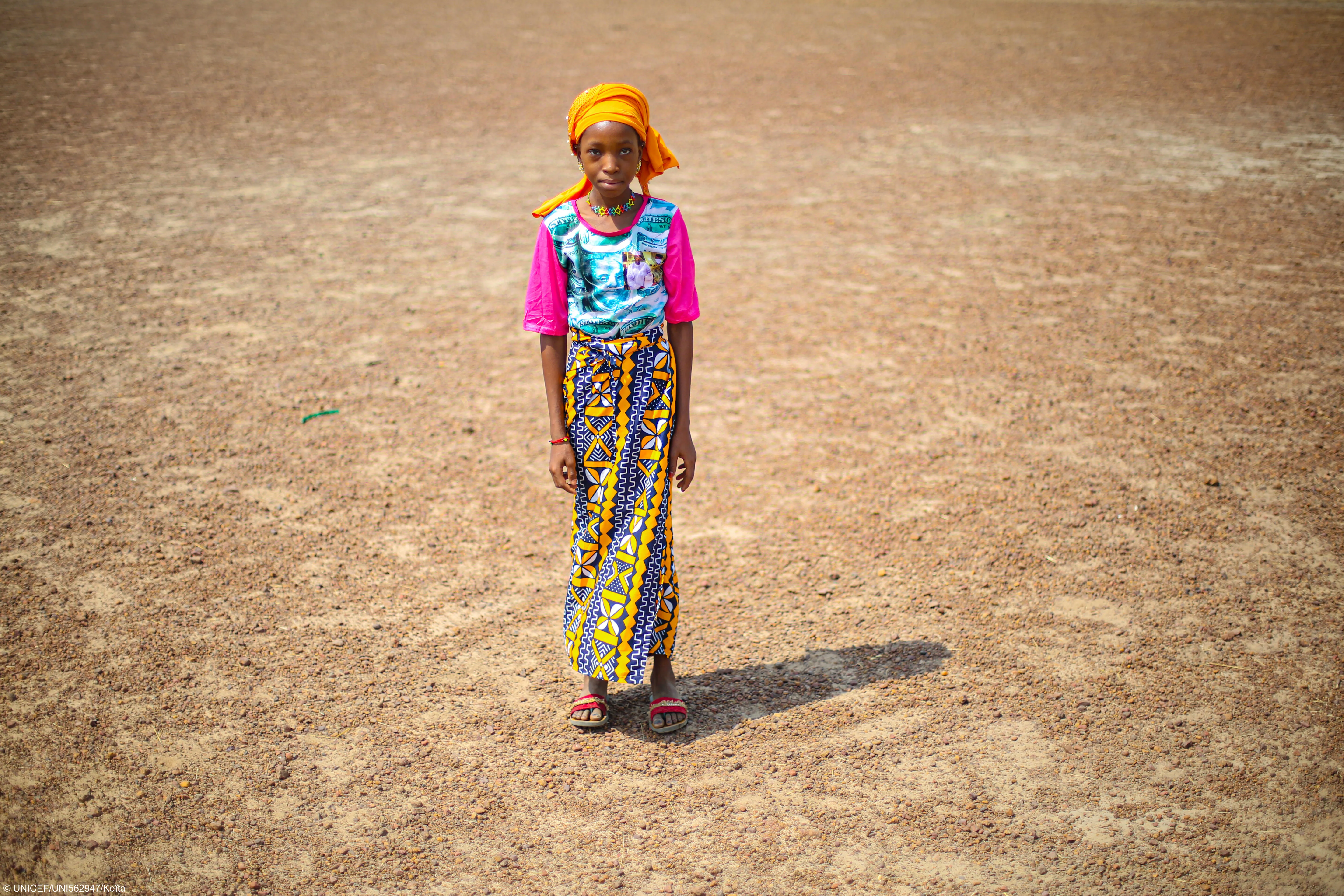 Une jeune fille se tient debout sur une terre aride, portant un chandail rose, une jupe à motifs, un foulard orange et des sandales.