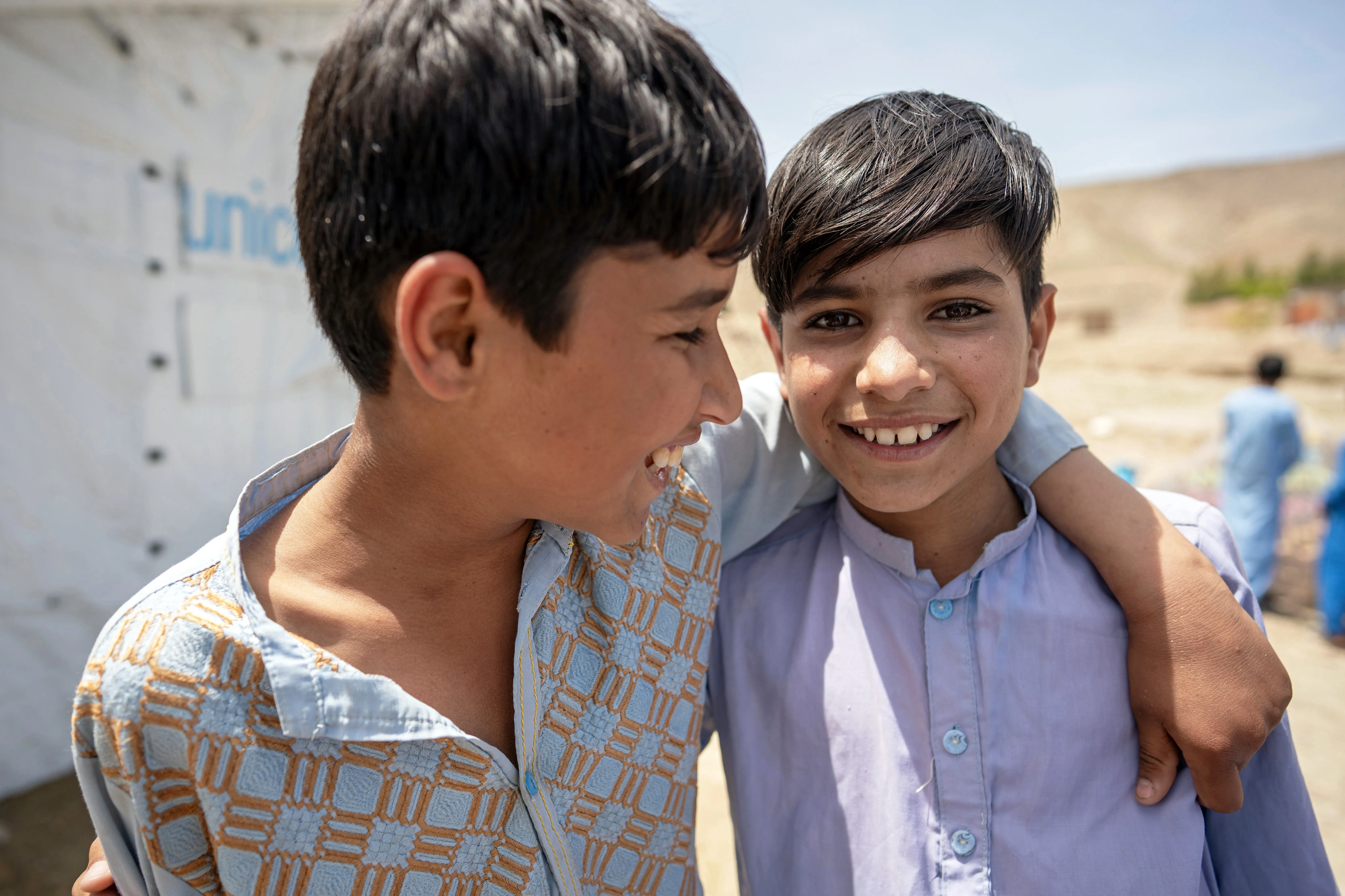 Two adolescent boys standing with their arms around each other’s shoulders and smiling outdoors in the sunshine, in front of a UNICEF tented shelter.