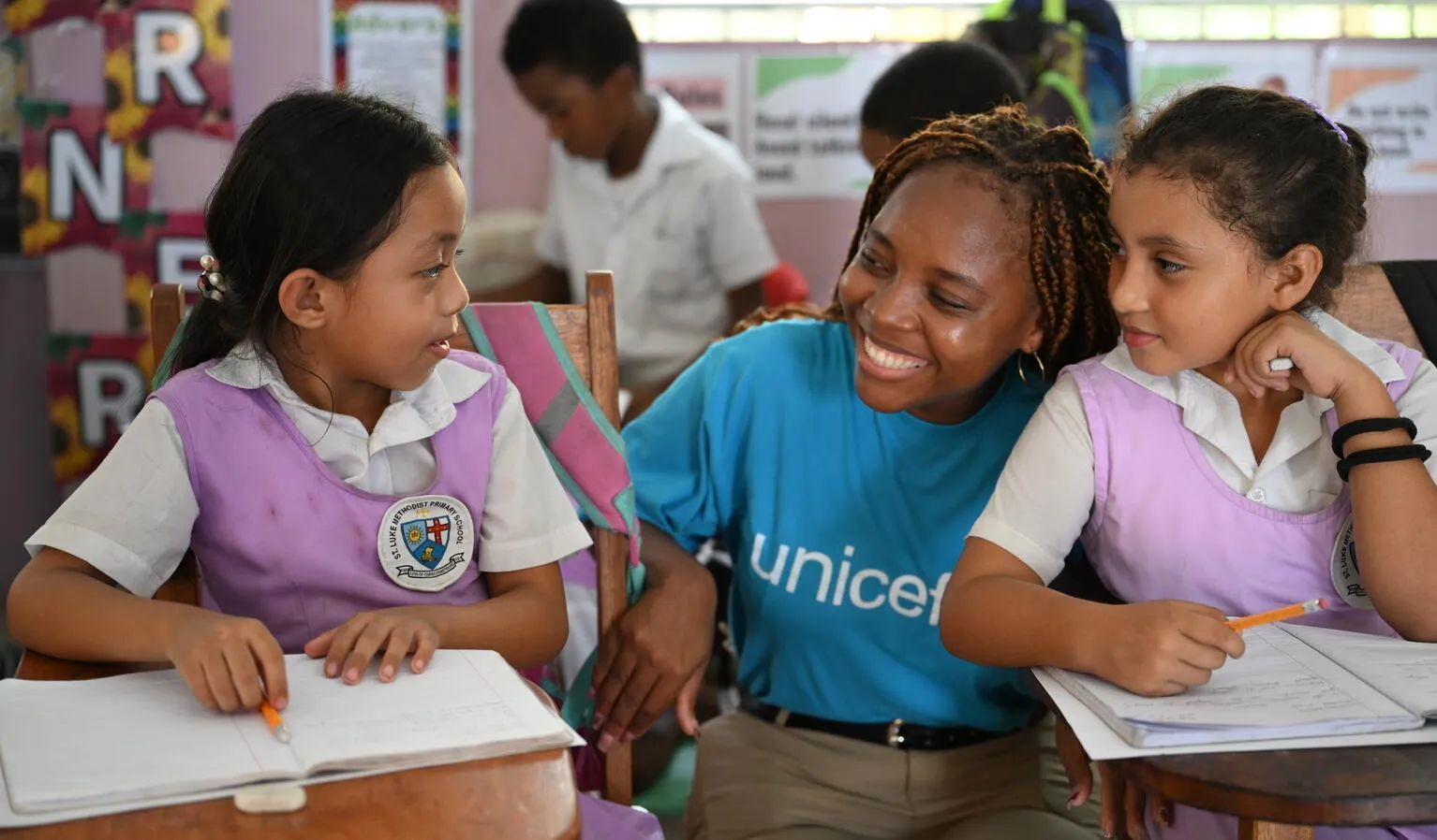 In a classroom, a woman wearing a blue UNICEF shirt crouch and smiles beside two girls sitting at their desk.