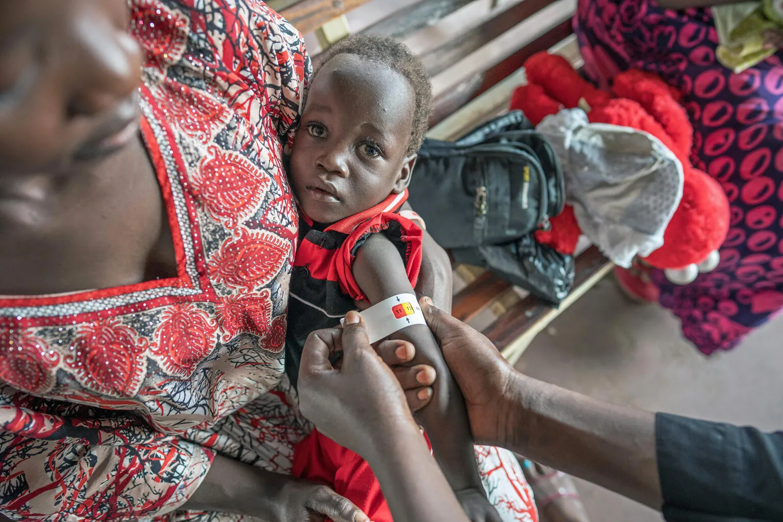 A caregiver holds a young child on her lap. The child wears a red and black outfit as a medical worker measures the child's upper arm with a MUAC tape to assess malnutrition.
