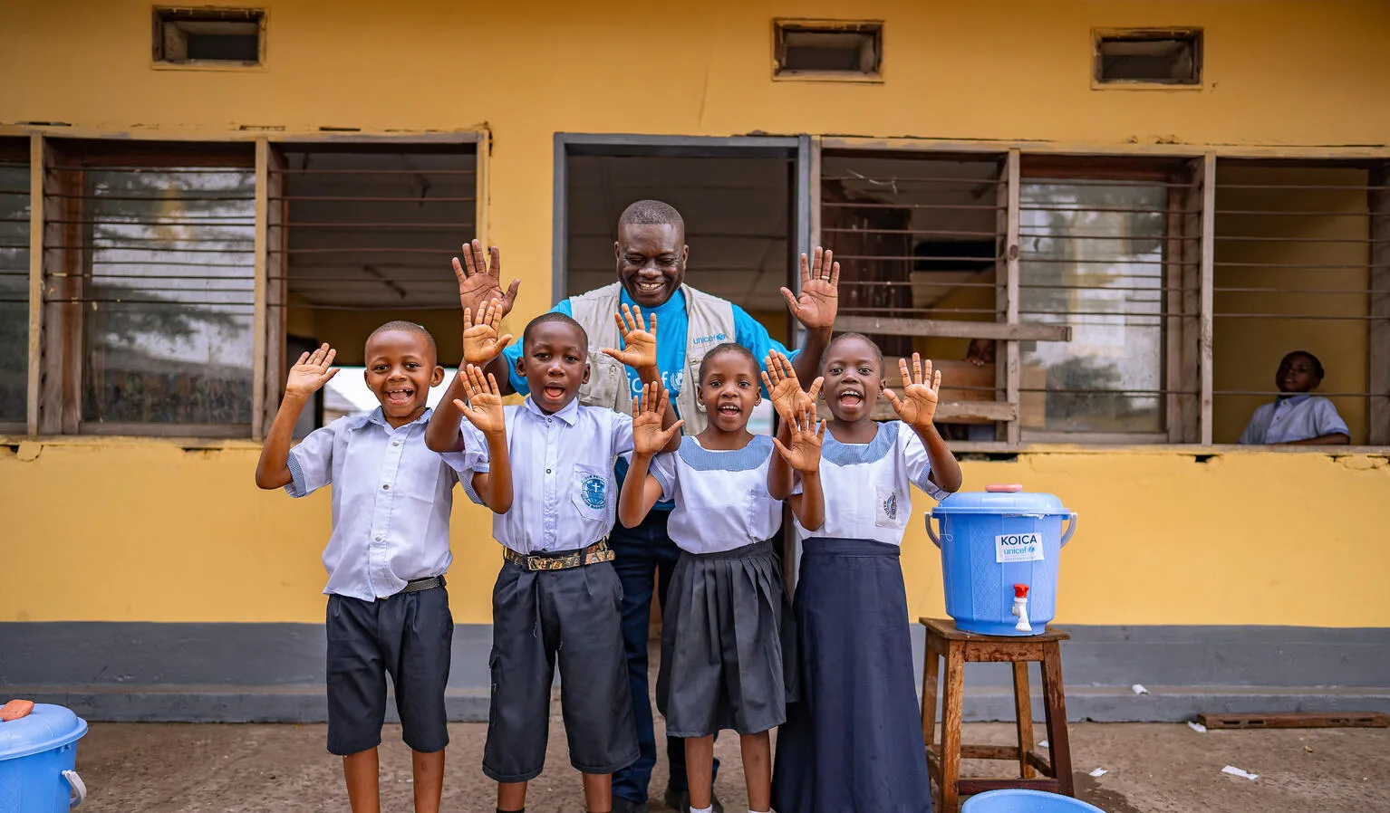 Un groupe d’enfants et un employé de l’UNICEF se tiennent à côté d’un seau d’eau, souriant à l’objectif tout en montrant leurs mains propres.