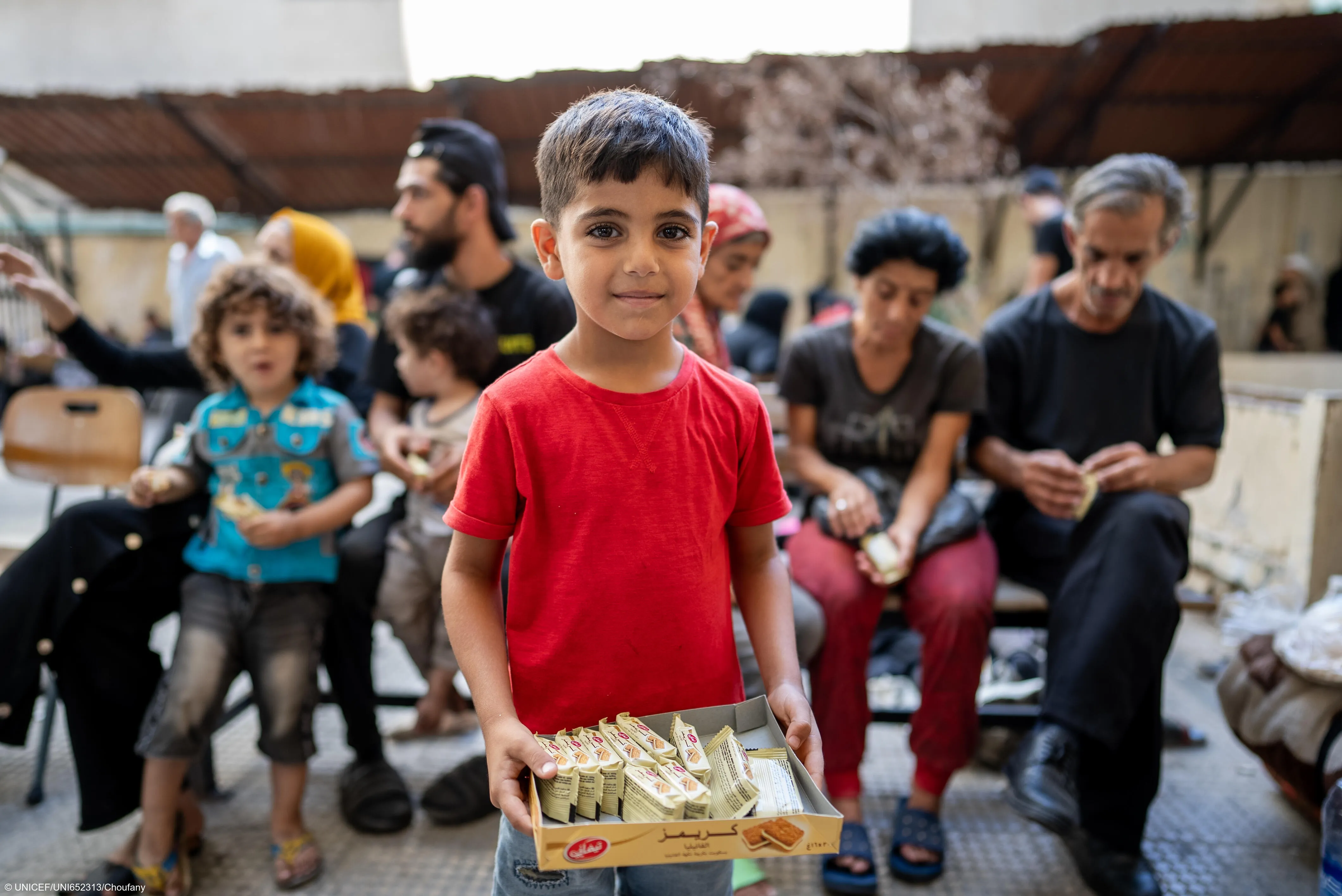 A boy in red shirt stands in front of people sitting together in Lebanon