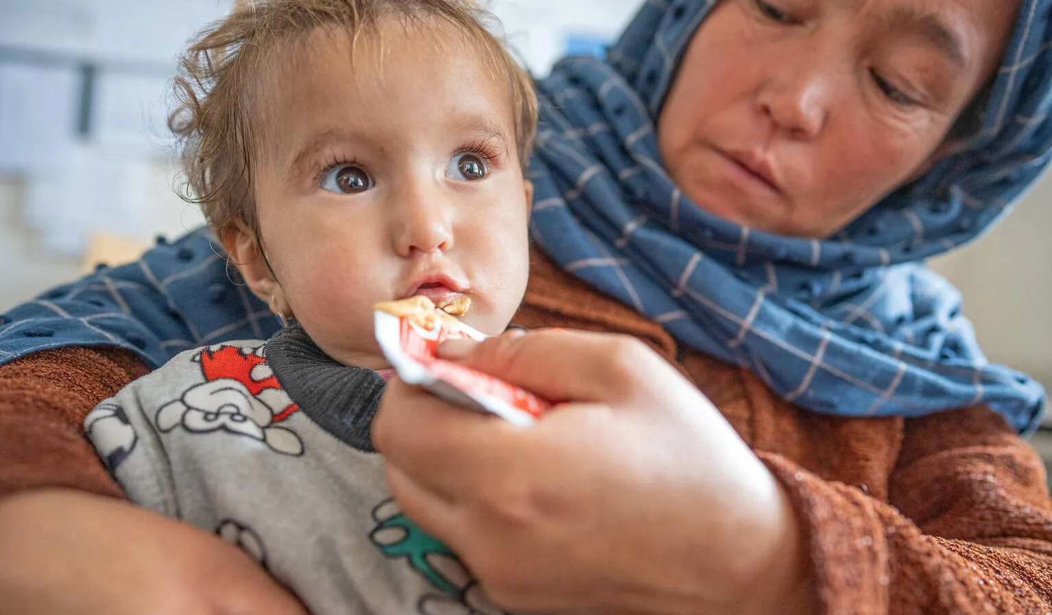 A baby eats a peanut paste out of a packet held by a caregiver in a blue headscarf.