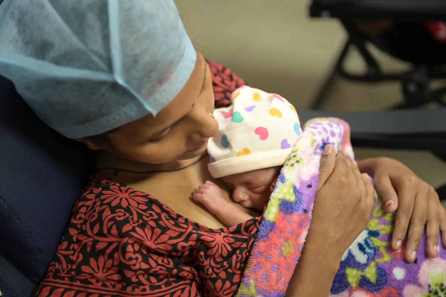 A mother wearing a surgical cap lovingly cradles her newborn, wrapped in a vibrant blanket. The baby wears a hat with colorful hearts, exuding warmth and care.