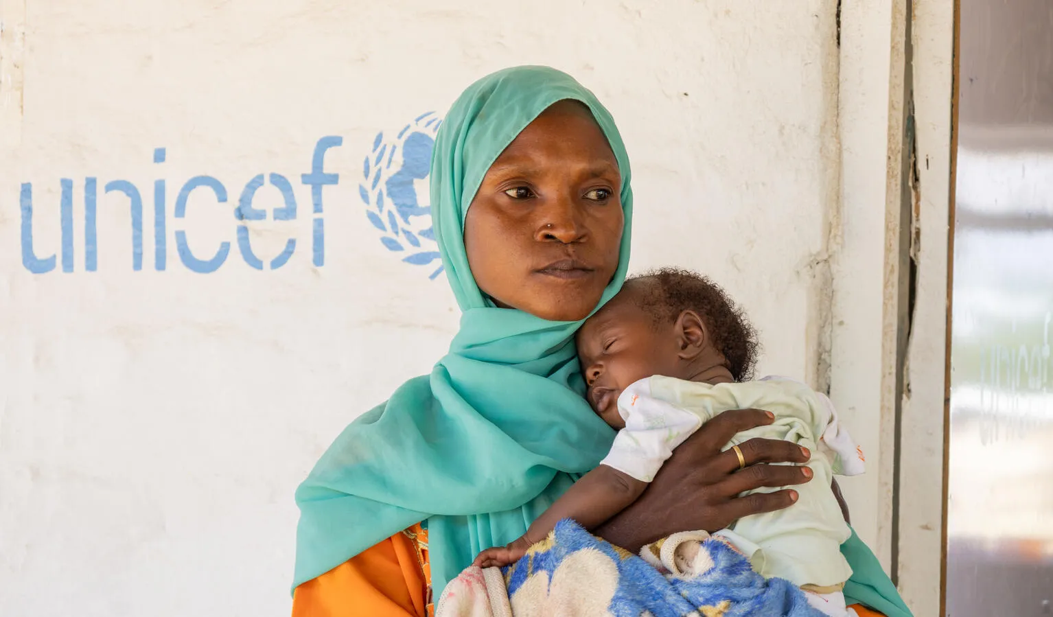 Une femme portant un foulard vert tient son bébé devant un mur orné du logo de l’UNICEF.