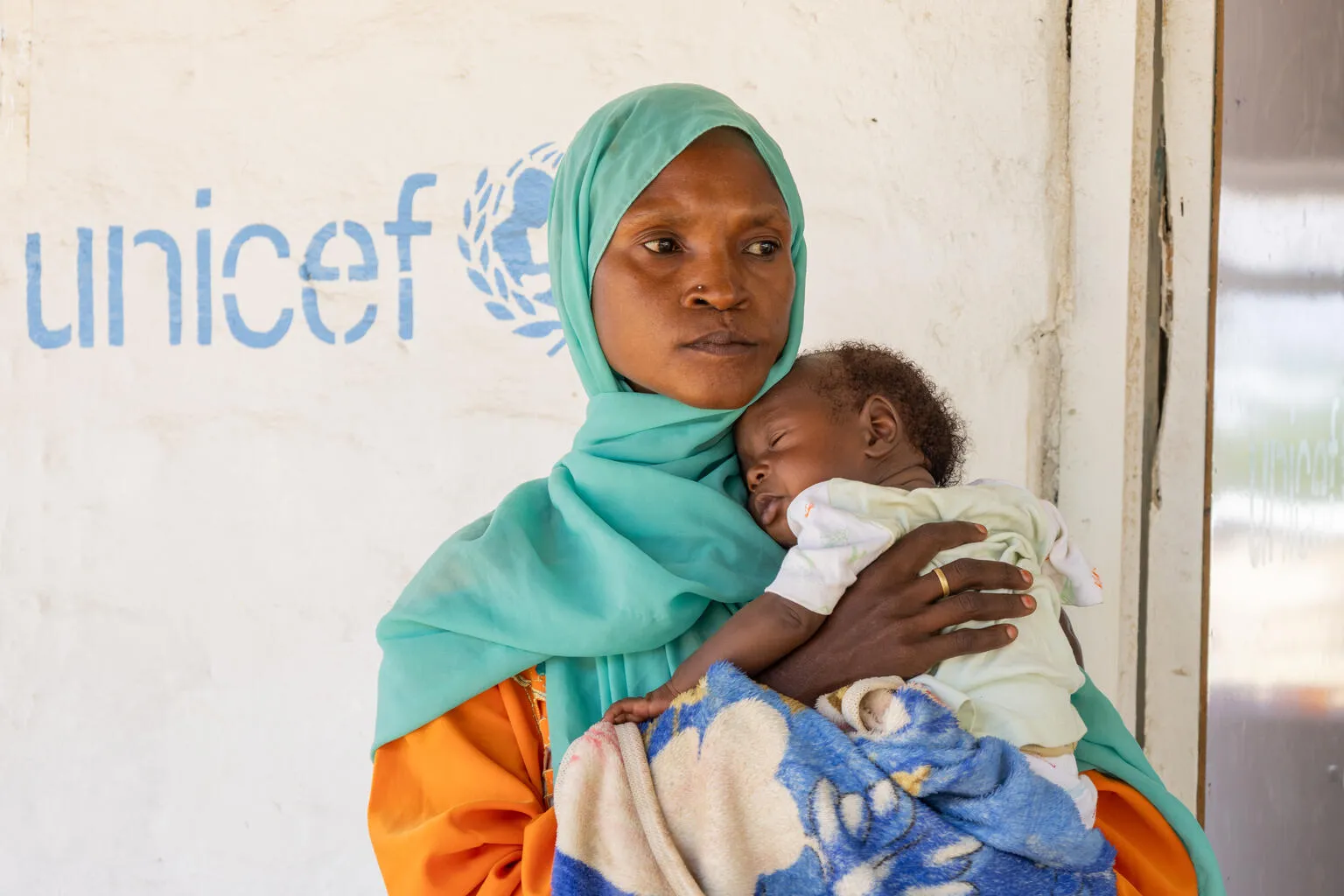  A woman in a green headscarf holds her baby in front of a wall with the UNICEF logo on it.