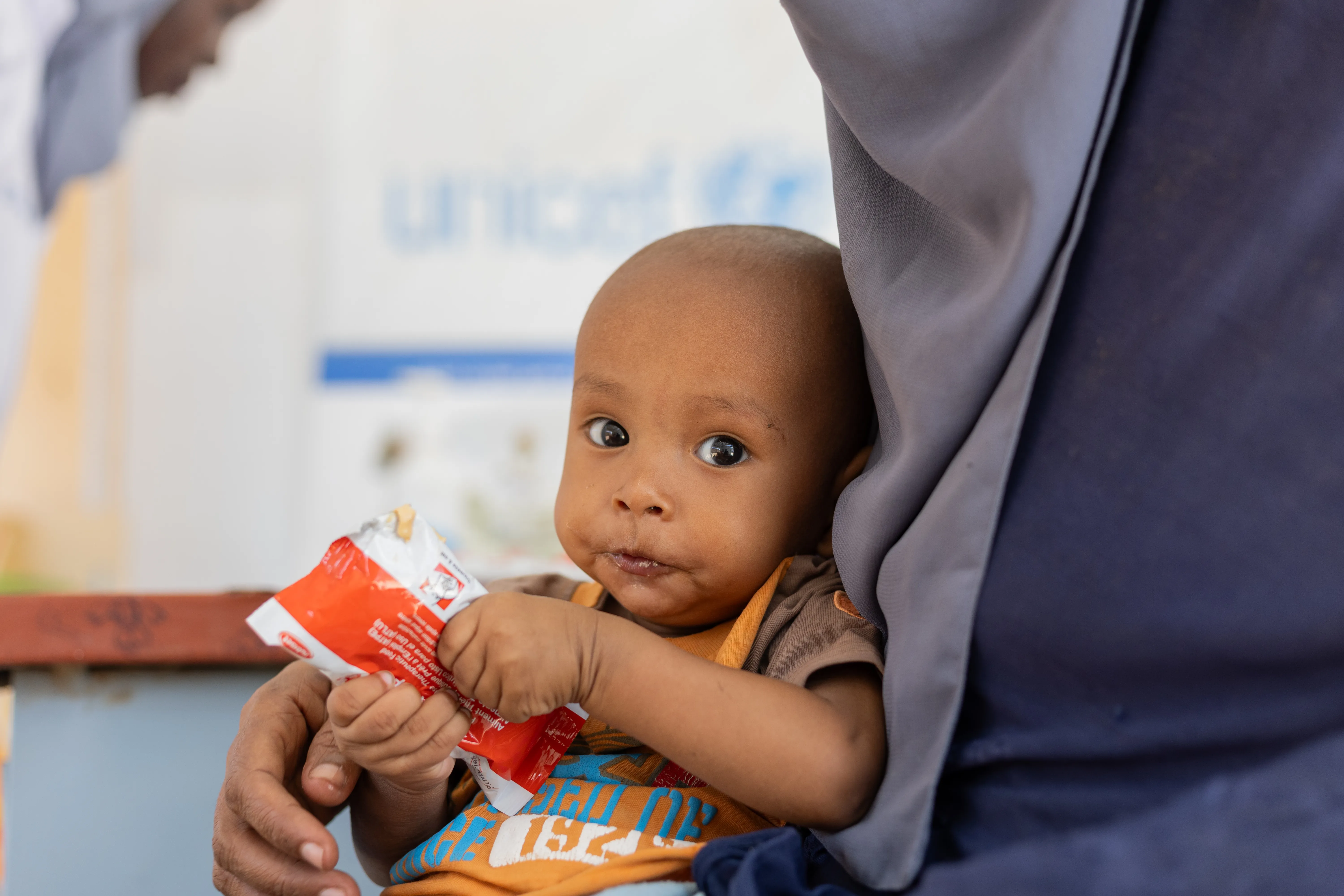A child sits in their mother's lap as they hold a packet of RUTF in Sudan