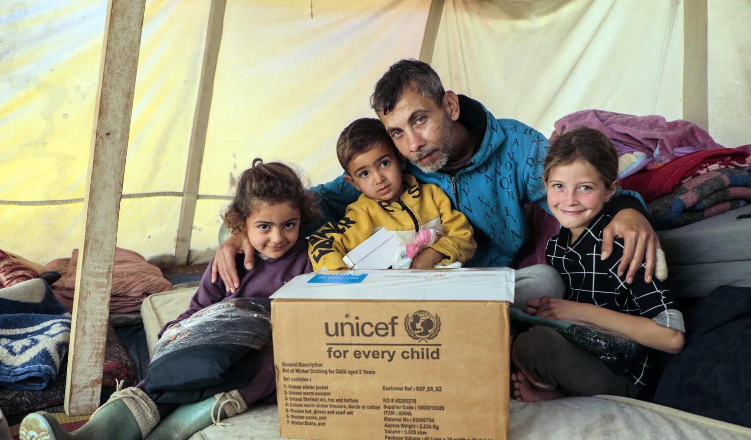 In an emergency tent, three children sit on the floor with their father who has his arms around them. In front of them is a UNICEF supply box.