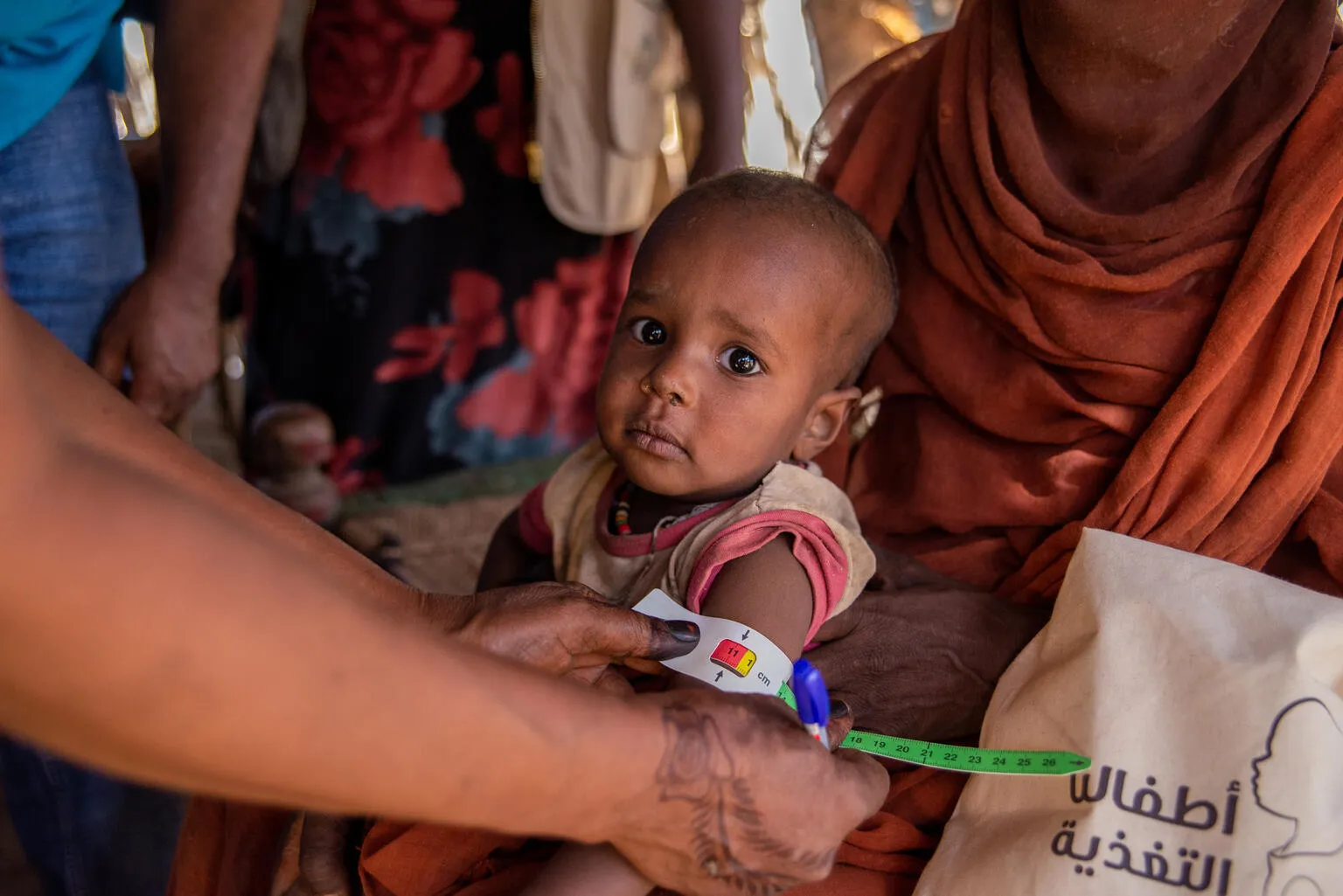 A health worker measures the upper arm of a young child sitting on a woman's lap with a colored band. The setting appears to be an aid center.
