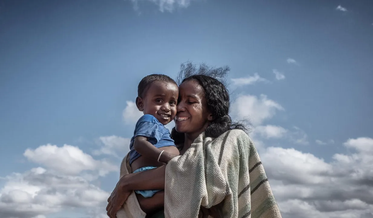 Une femme qui sourit tient dans ses bras un jeune enfant à l’extérieur, sous un ciel partiellement nuageux.
