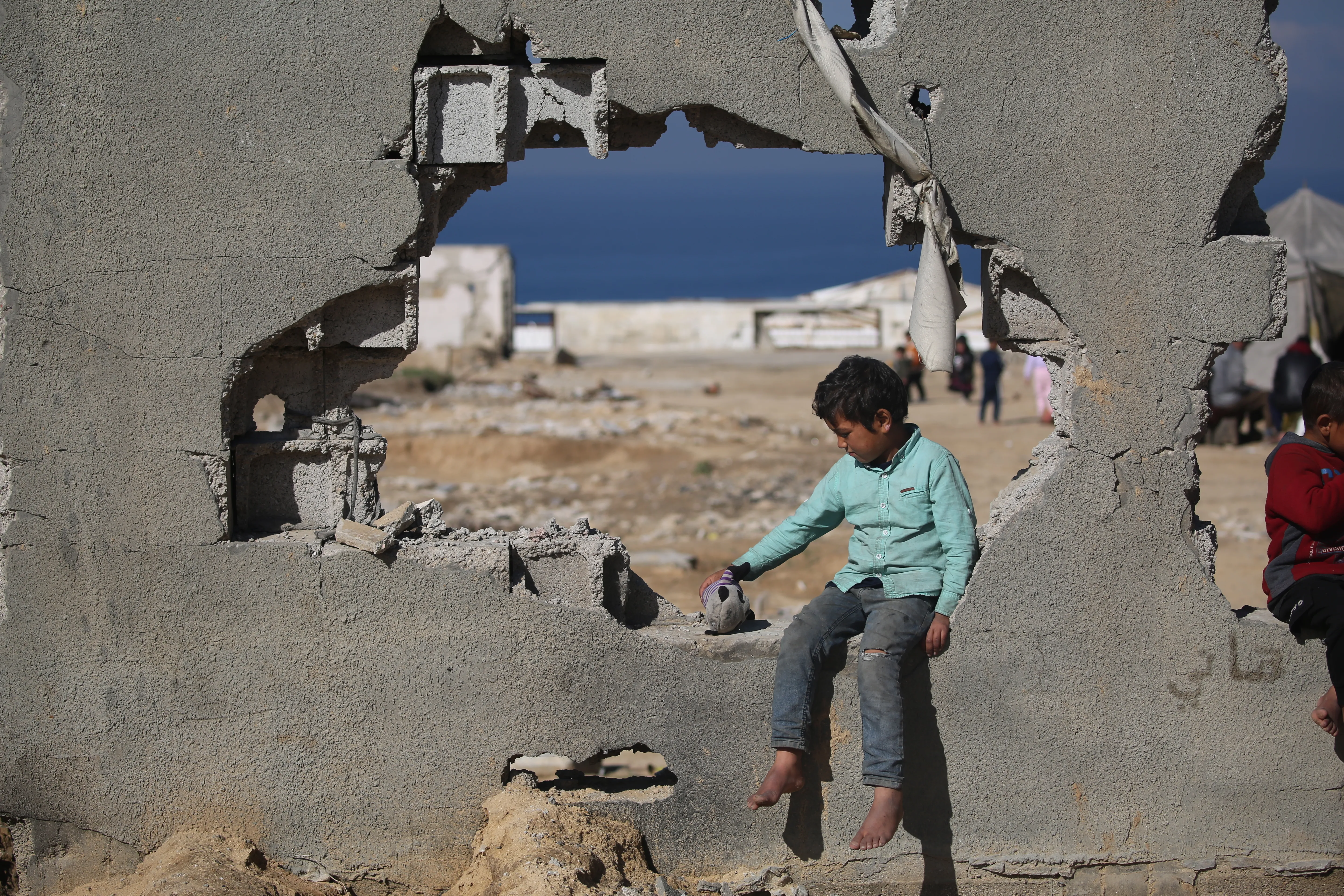 Barefoot boy sitting in a hole in a damaged concrete wall.