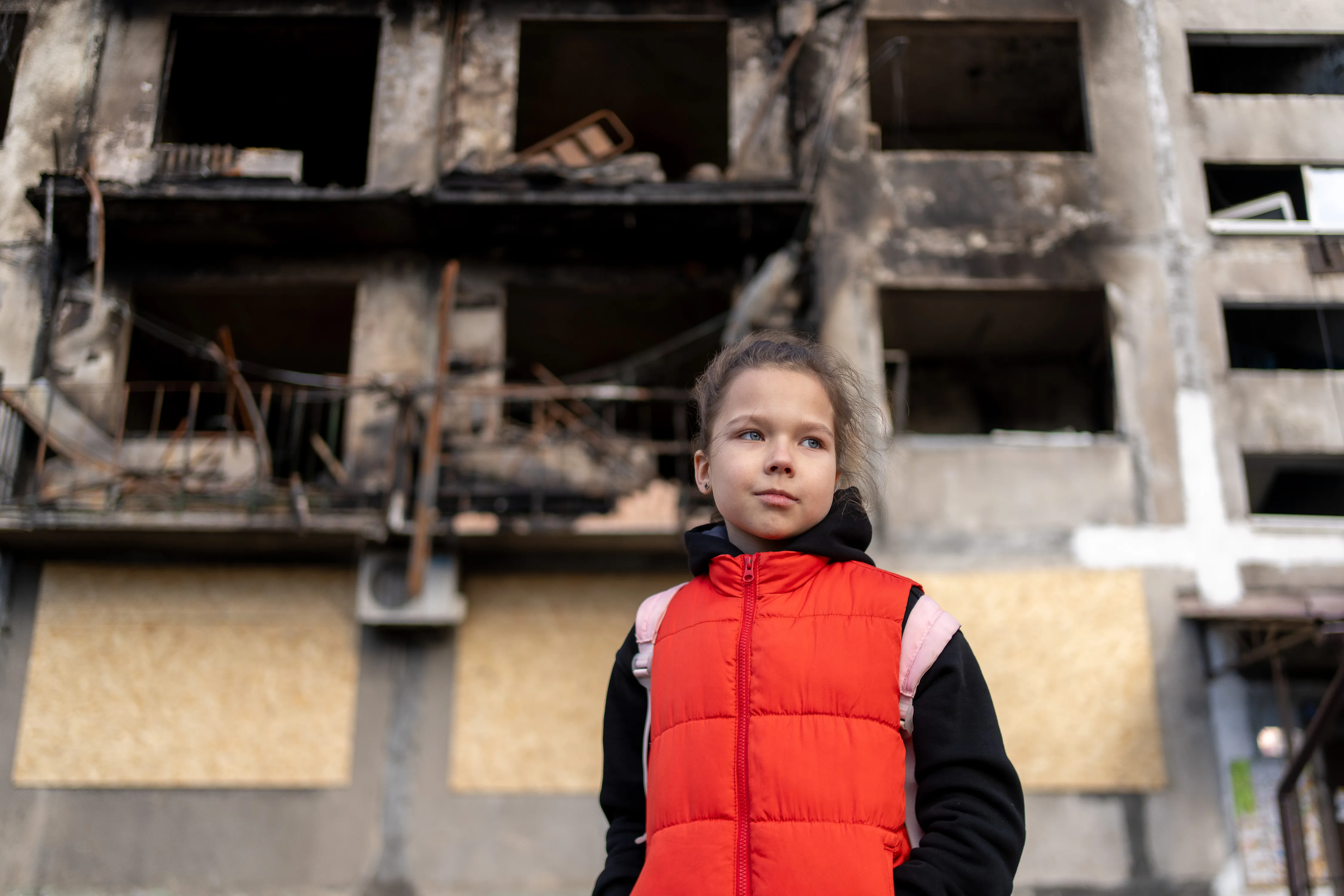Girl wearing a red vest and backpack standing in front of a fire-damaged apartment building.