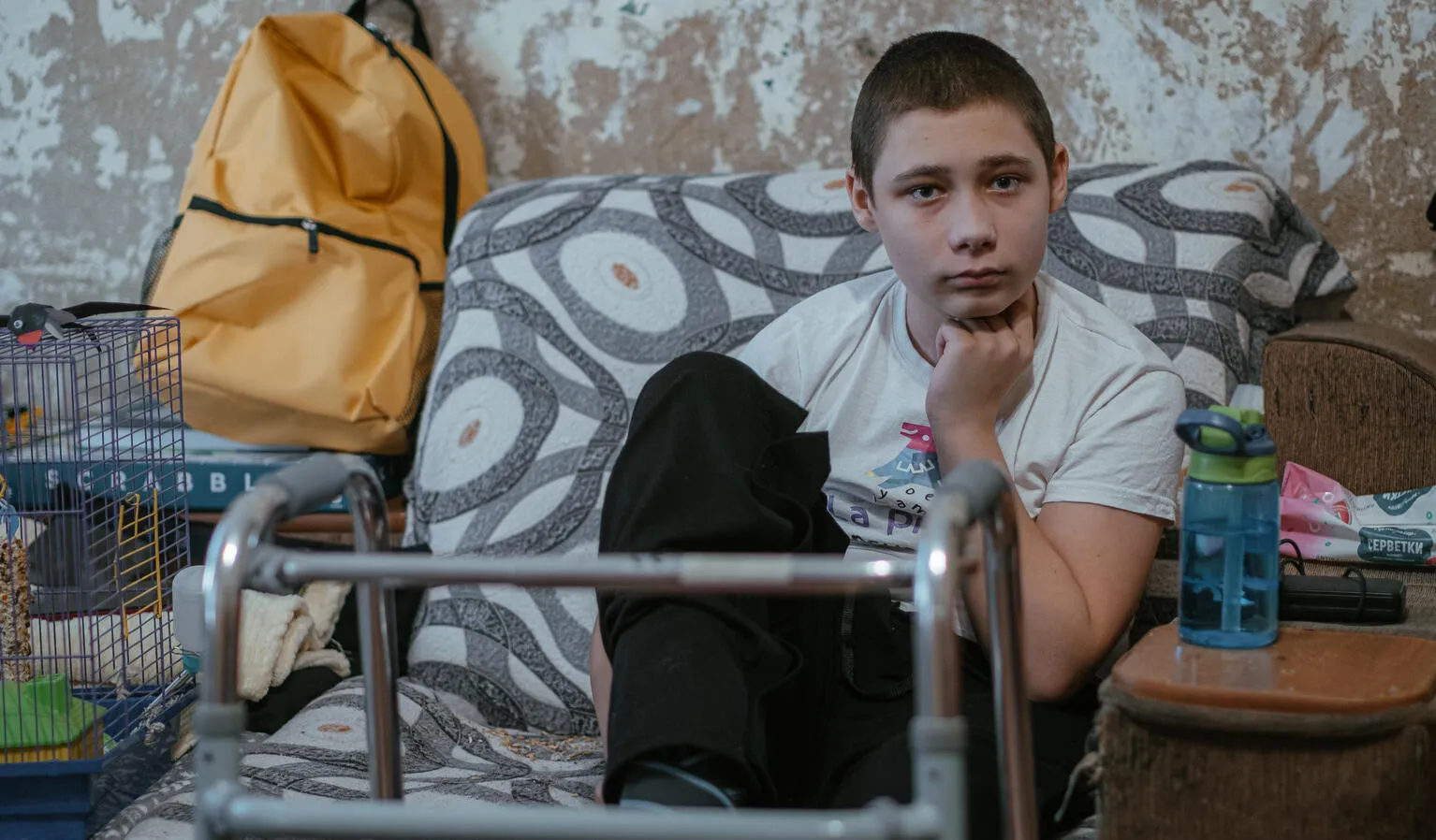 A boy sits on a patterned sofa, looking thoughtful. A walker is in front of him, a yellow backpack and hamster cage are nearby, and a bottle is on a table.