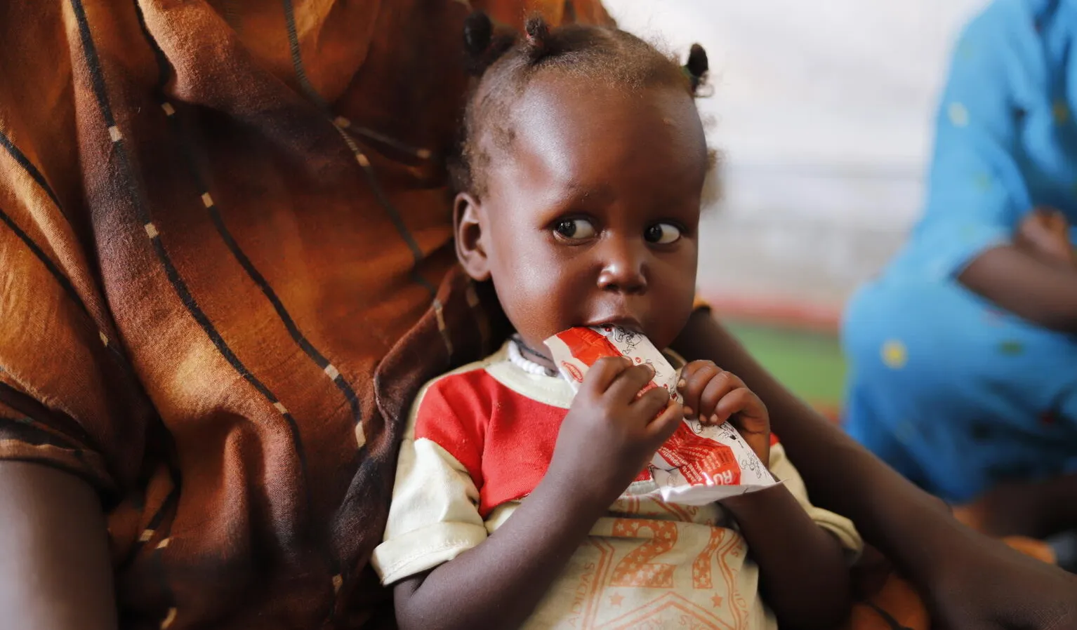 A small child sits on an adult's lap, holding a packet of nutritional paste to their mouth. The child's expression is thoughtful. The adult wears a patterned brown garment.
