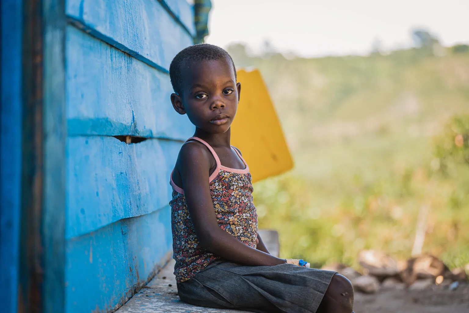 A young child sits on a ledge beside a bright blue wooden wall, gazing thoughtfully at the camera. The background features a blurred, green landscape