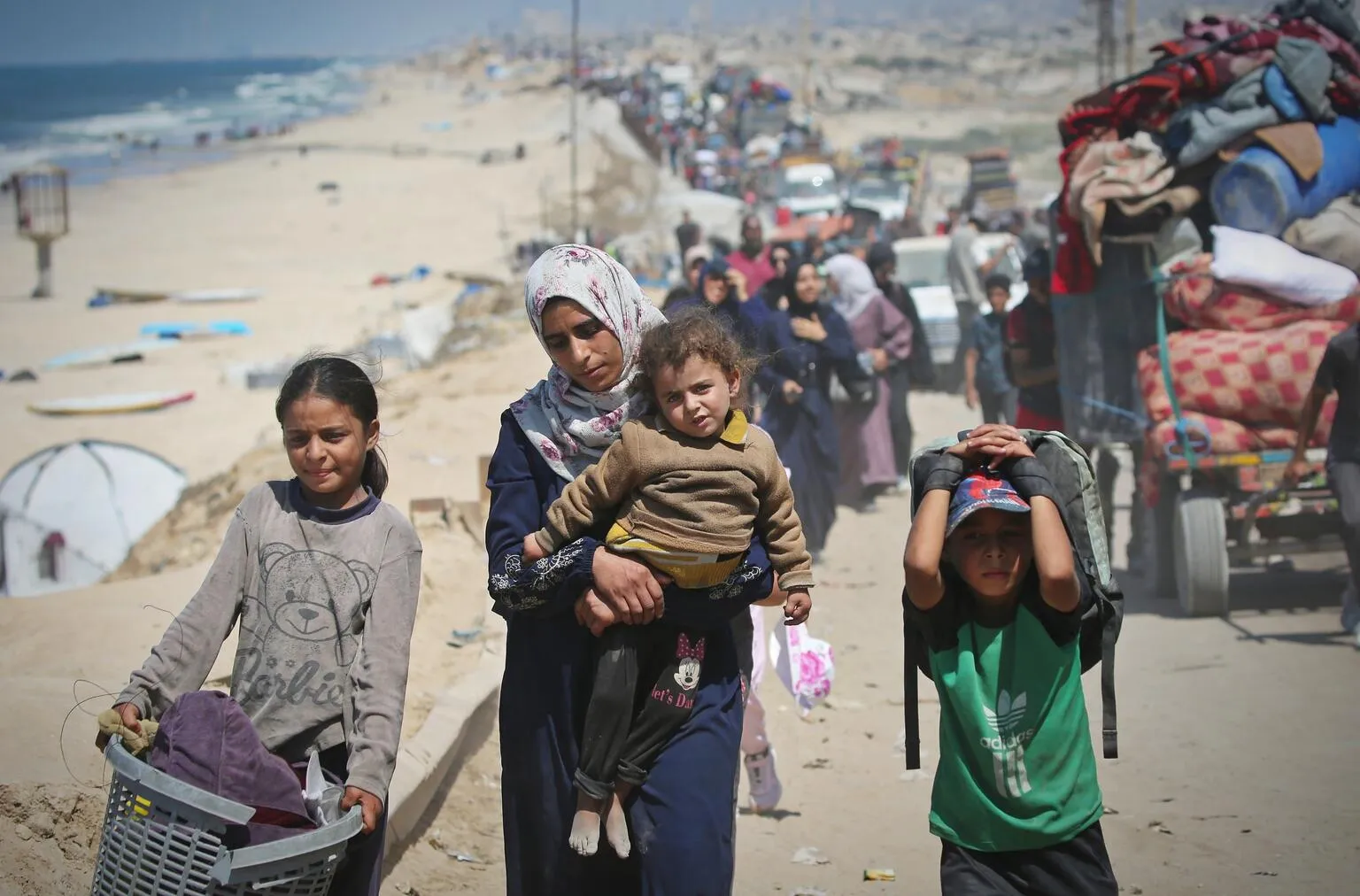 Une femme portant un enfant, une fille tenant un panier et un garçon portant un sac à dos marchent sur une route poussiéreuse avec d’autres personnes.