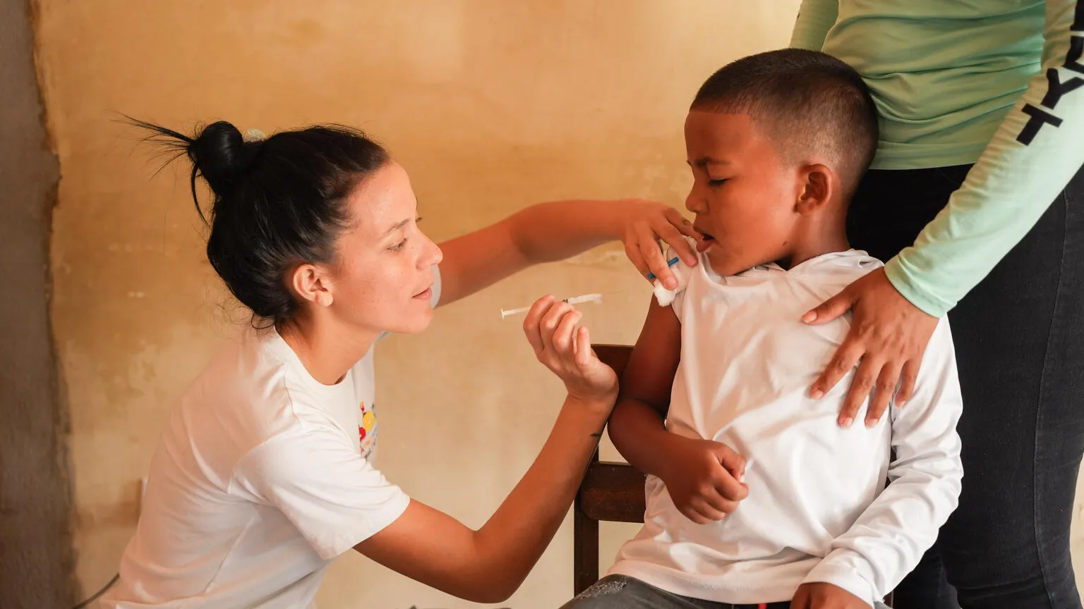 A healthcare worker administers a vaccine to a young boy sitting calmly, with an adult’s supportive hand on his shoulder. 