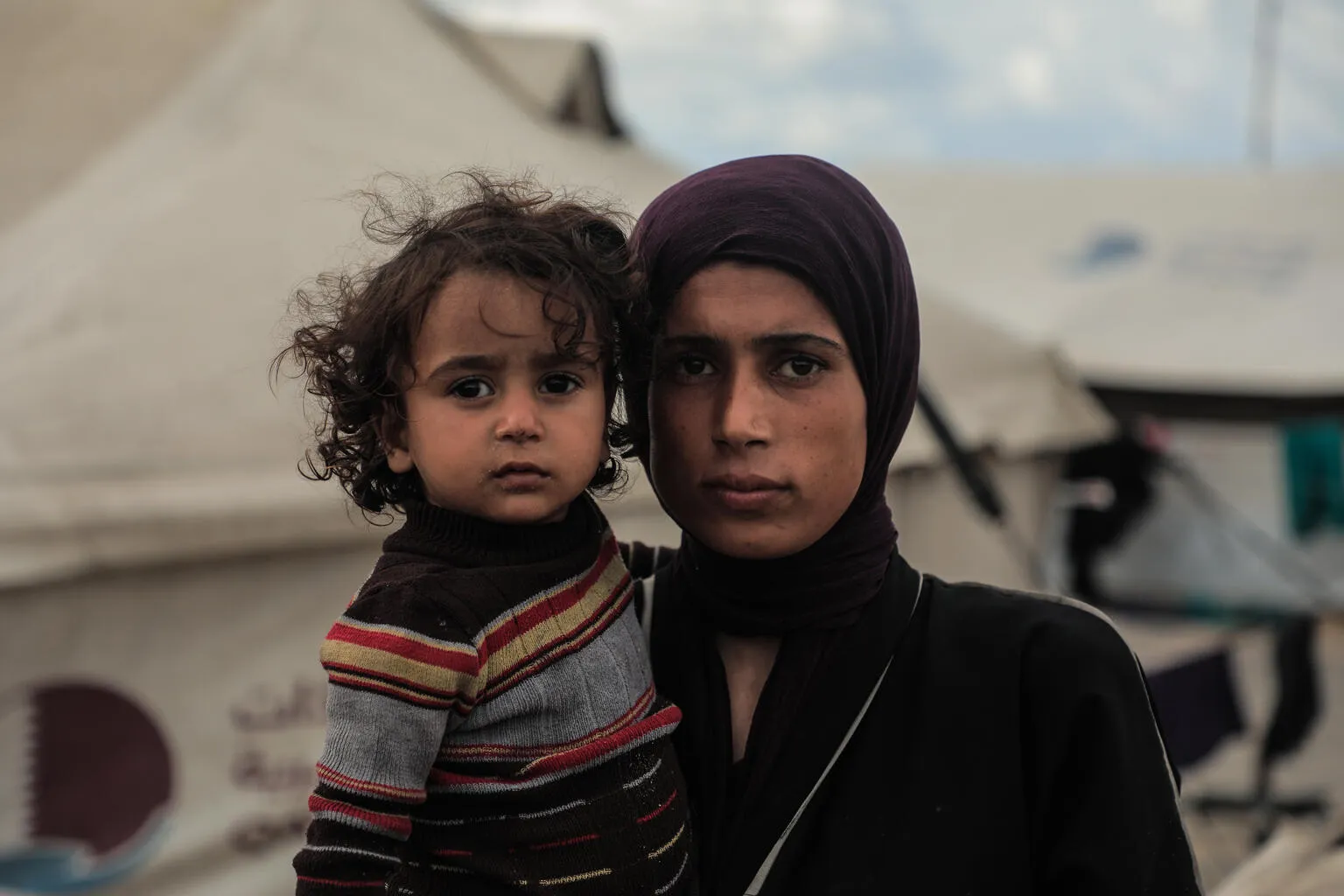 A woman in a purple headscarf and dark clothing holds a young child in a striped sweater. They stand in front of tents, conveying a somber tone.