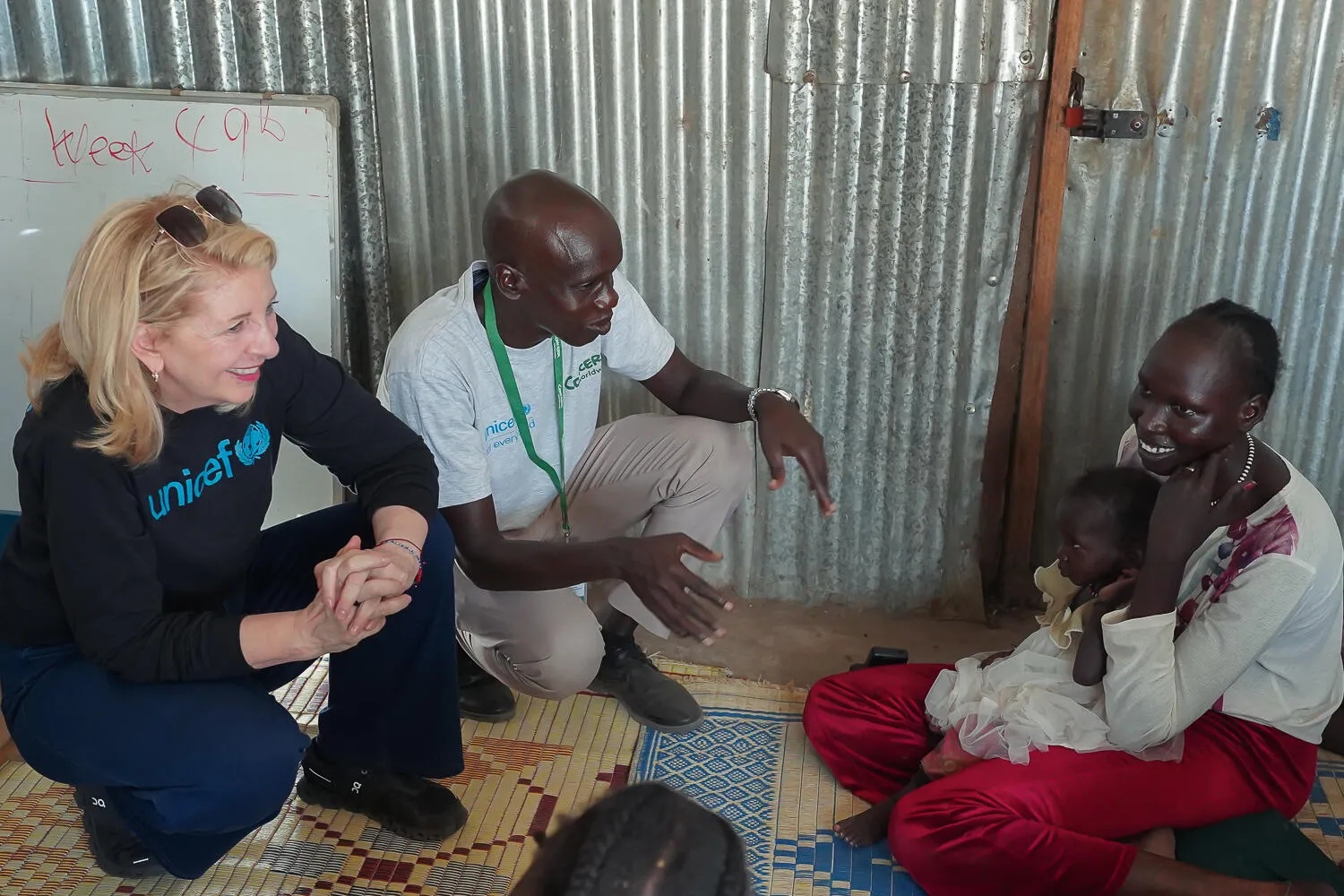 A woman in a UNICEF sweater and a man converse with a smiling woman holding a child, seated on mats in a metal-walled room, conveying warmth and hope.