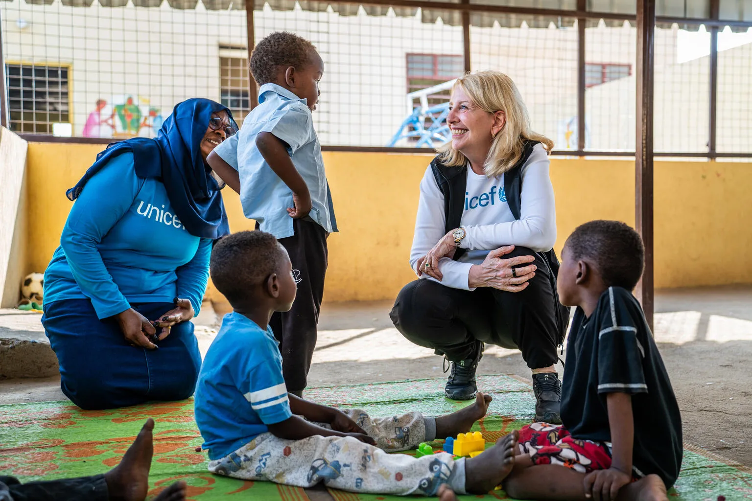 A woman in a UNICEF shirt smiles while kneeling in front of children in a play area. The warm atmosphere is supportive and engaging.