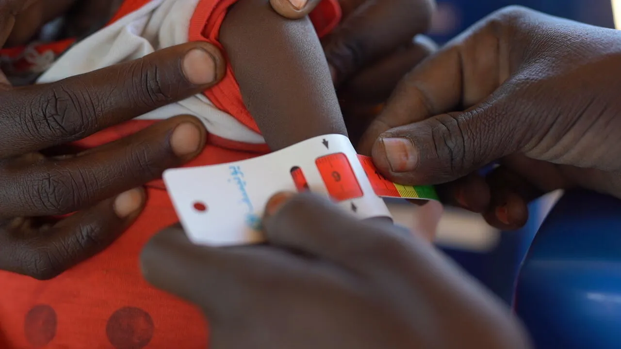 A child's arm is measured with a MUAC tape by two adults, wearing a red and white garment. The scene conveys care and urgency in a healthcare context.