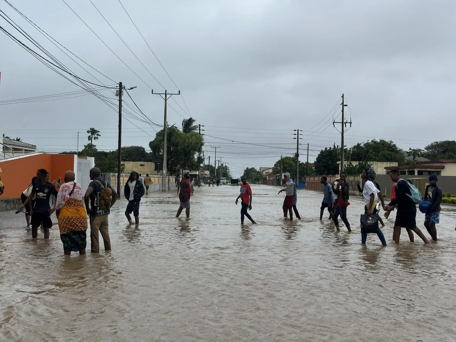 People walking through a flooded street under gray skies. The water covers the road, and they hold hands, conveying a sense of solidarity and resilience.
