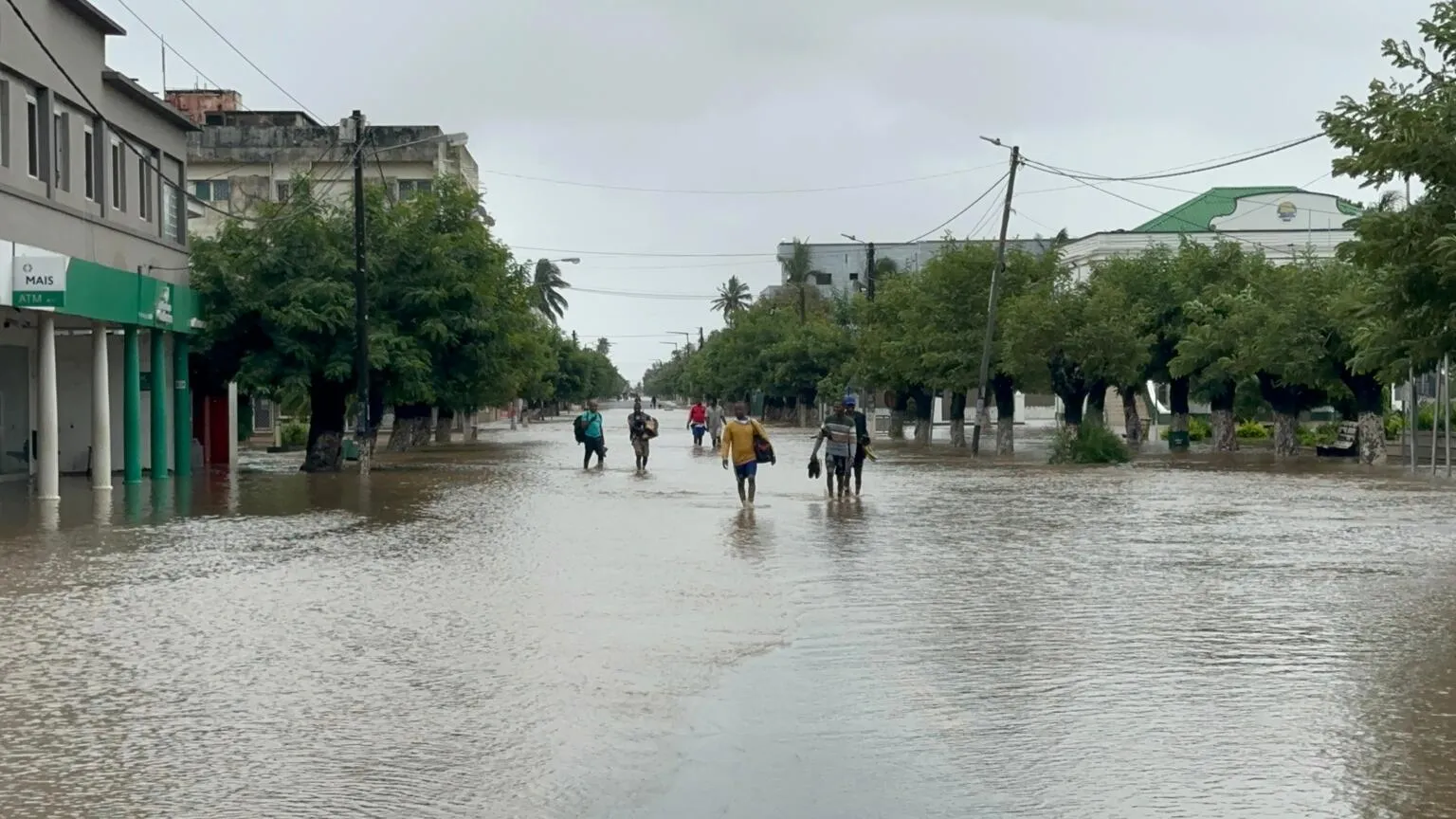 A flooded urban street with submerged road, people wading through knee-deep water, overcast skies, and trees lining the street, conveying a somber mood.
