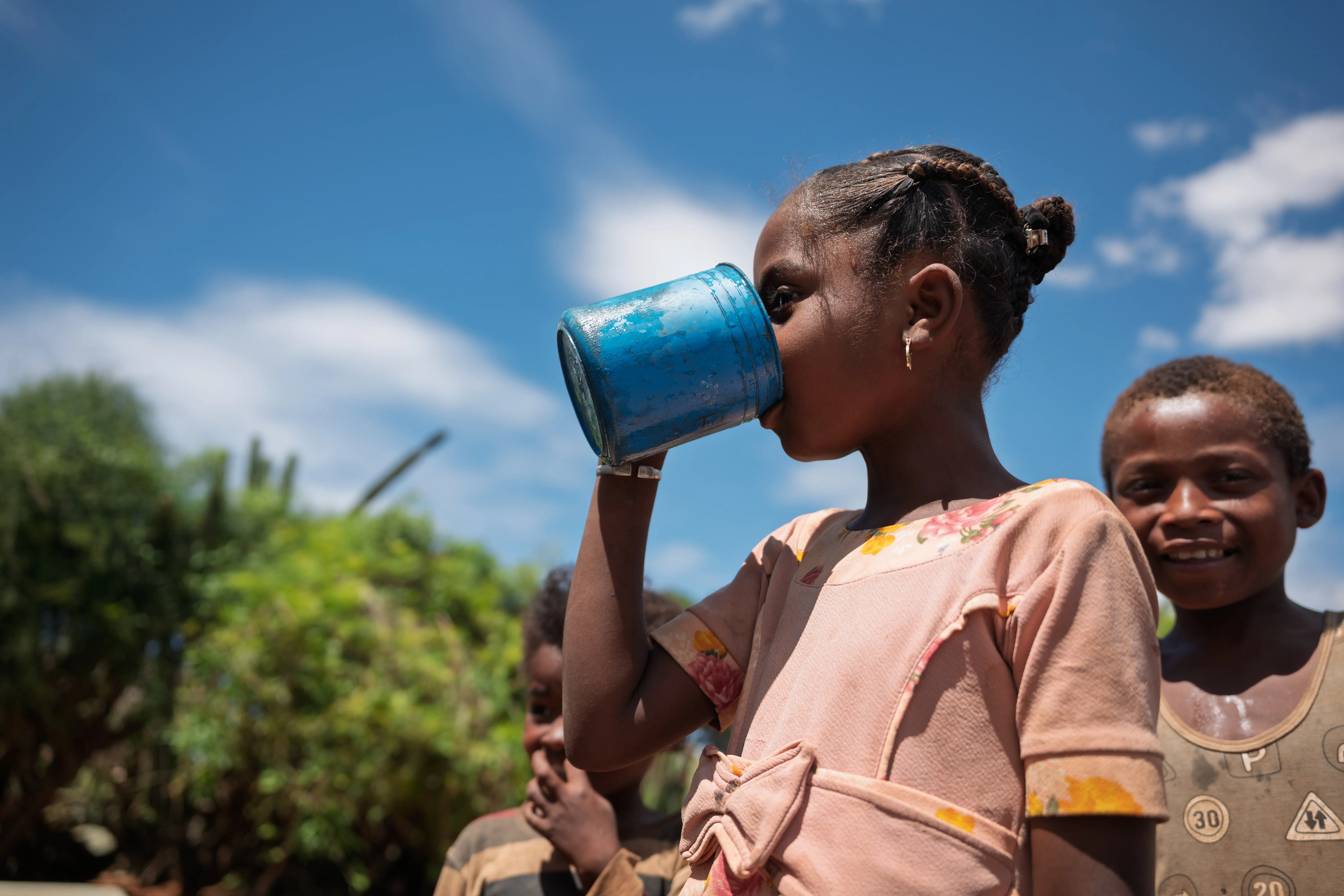 Girl drinking water from a metal cup, with other children nearby outdoors.
