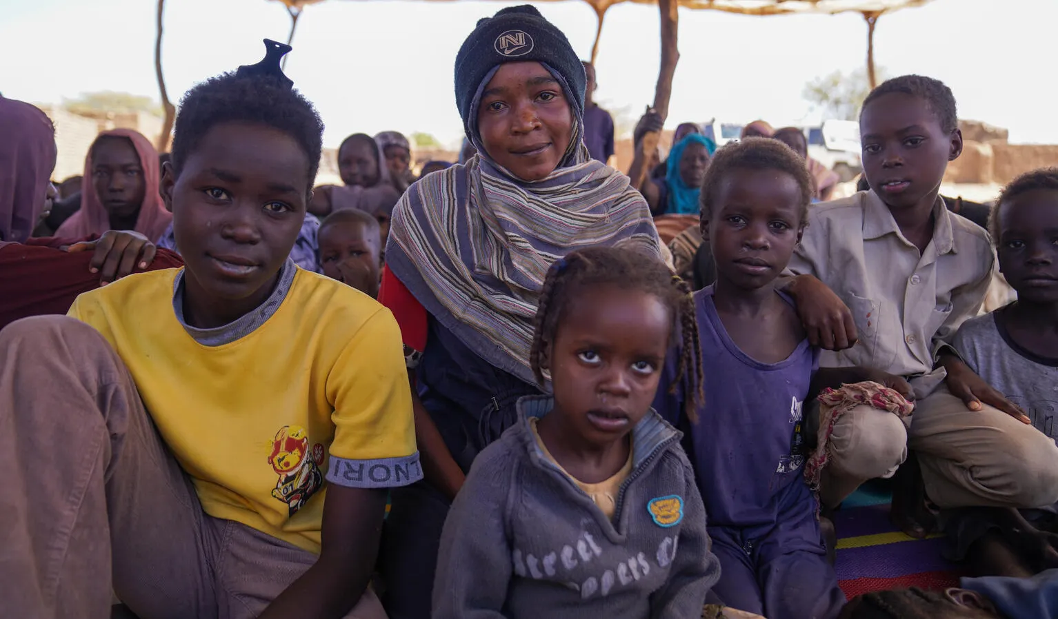 A group of children sit closely together under a makeshift tent. The scene conveys resilience and community.