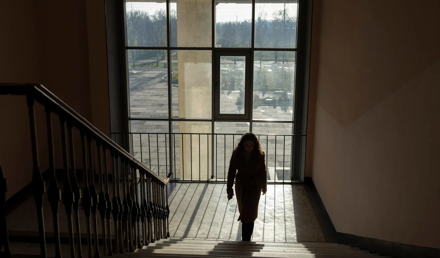 A person walks up a dimly lit staircase, silhouetted against a large window that reveals an overcast outdoor scene. The mood is quiet and contemplative.