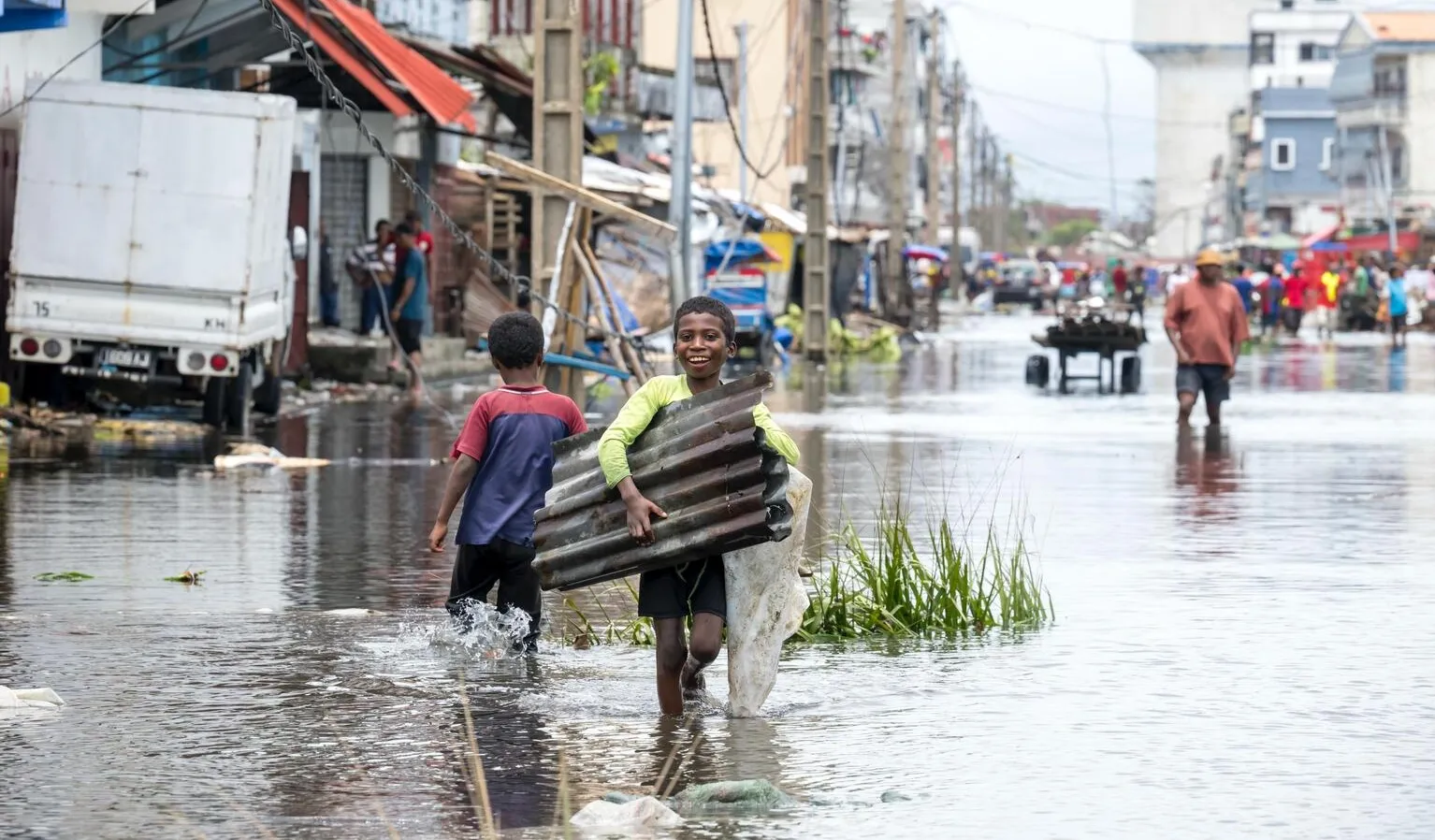 Two children navigate a flooded street, one carrying corrugated metal, both appearing determined. The scene is bustling with people amid submerged cars and buildings.