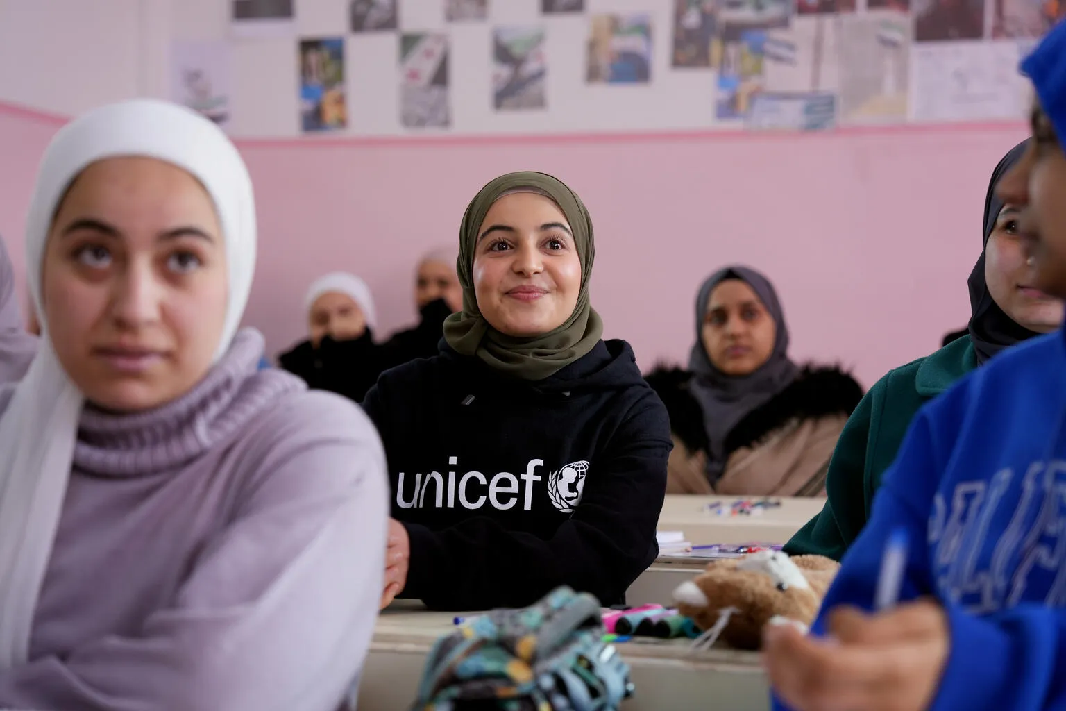 In a classroom with pink walls, a smiling girl wearing a hijab and a UNICEF shirt sits among attentive peers. The atmosphere is warm and positive.