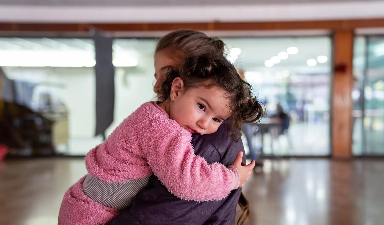 A mother holds a toddler wearing pink clothes in her arms in a brightly lit room. 