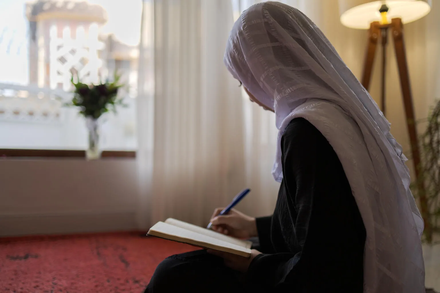 A girl in a white hijab is writing in a notebook, seated on a carpet. 