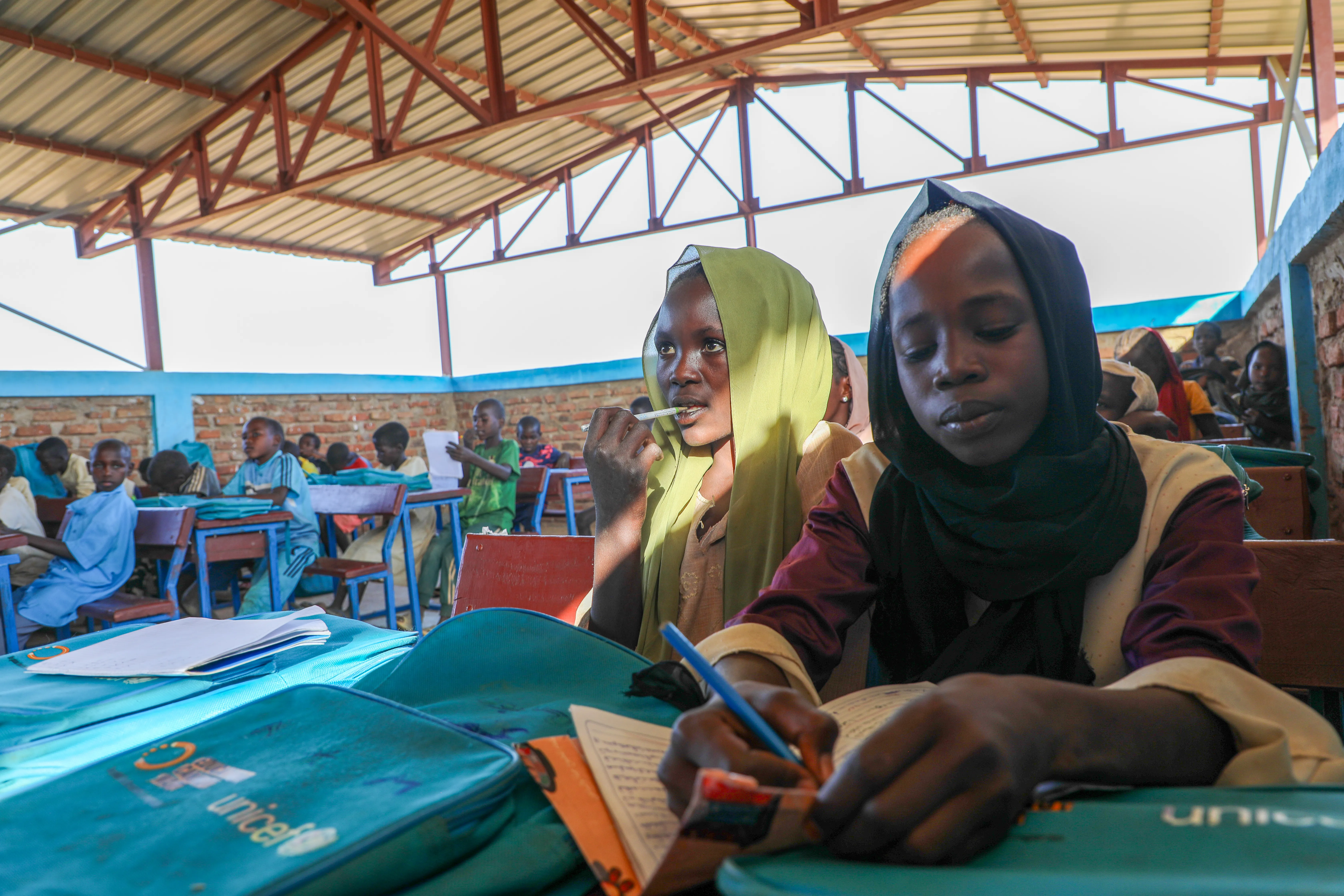 Girls in headscarves writing in notebooks at desks in an open-air classroom.
