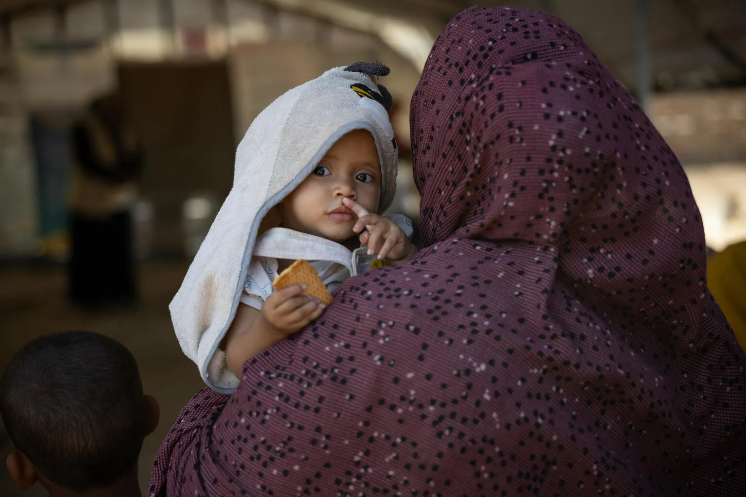 A child with a towel hood is held by a person in a patterned cloak, gazing intently and holding a piece of bread. 