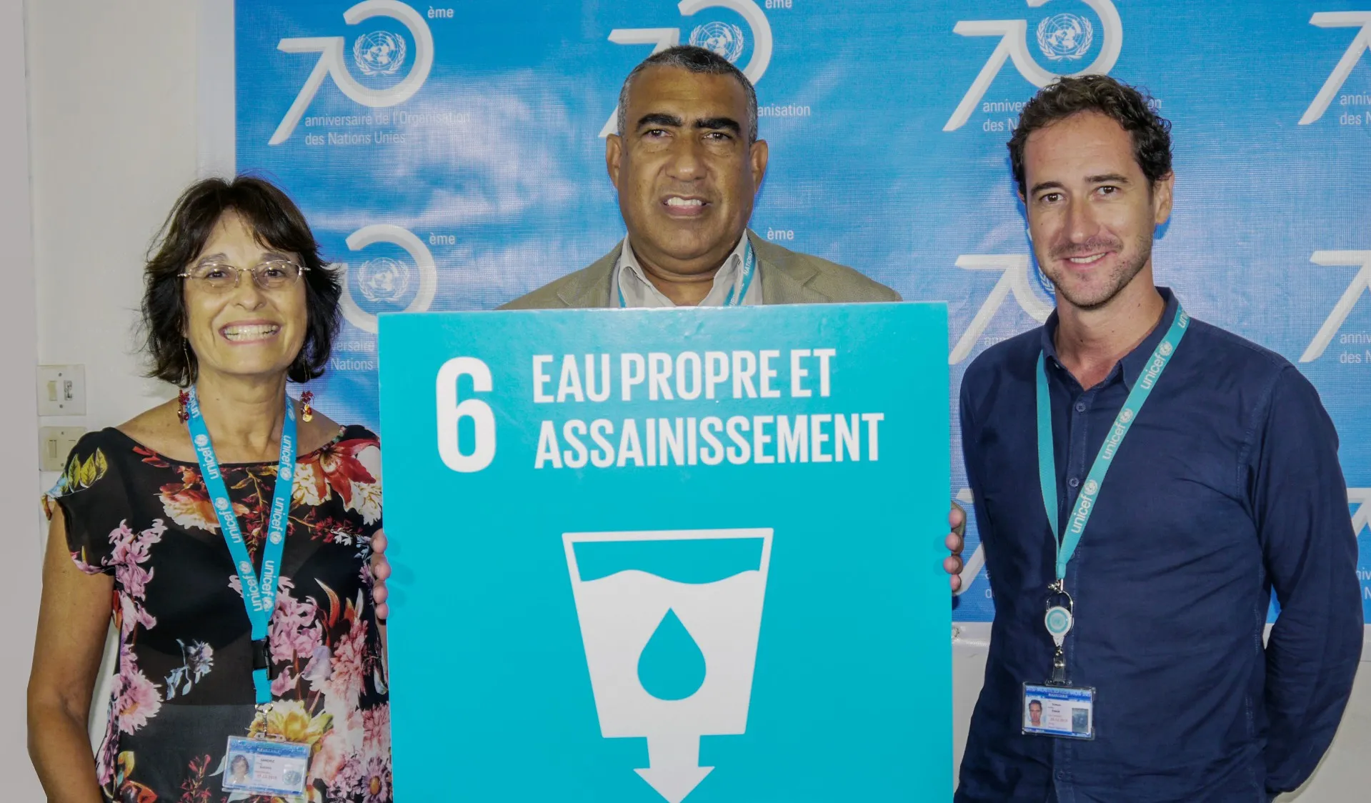 Three people smiling, standing in front of a blue banner, holding a sign that reads "Eau Propre et Assainissement" with the SDG 6 sign that has a water symbol.