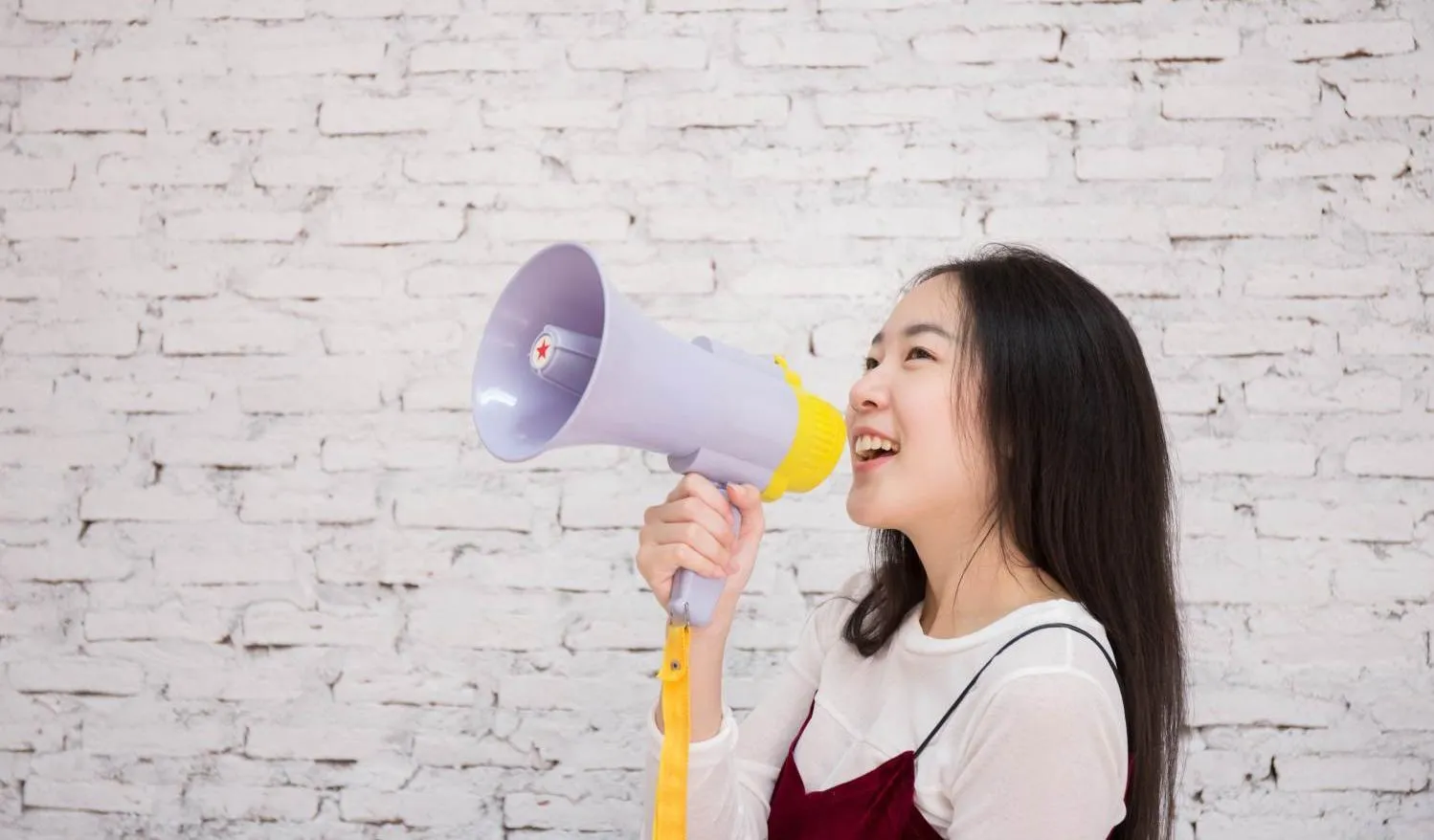 A young girl, standing alone in front of a white wall, smiles while holding up a megaphone.